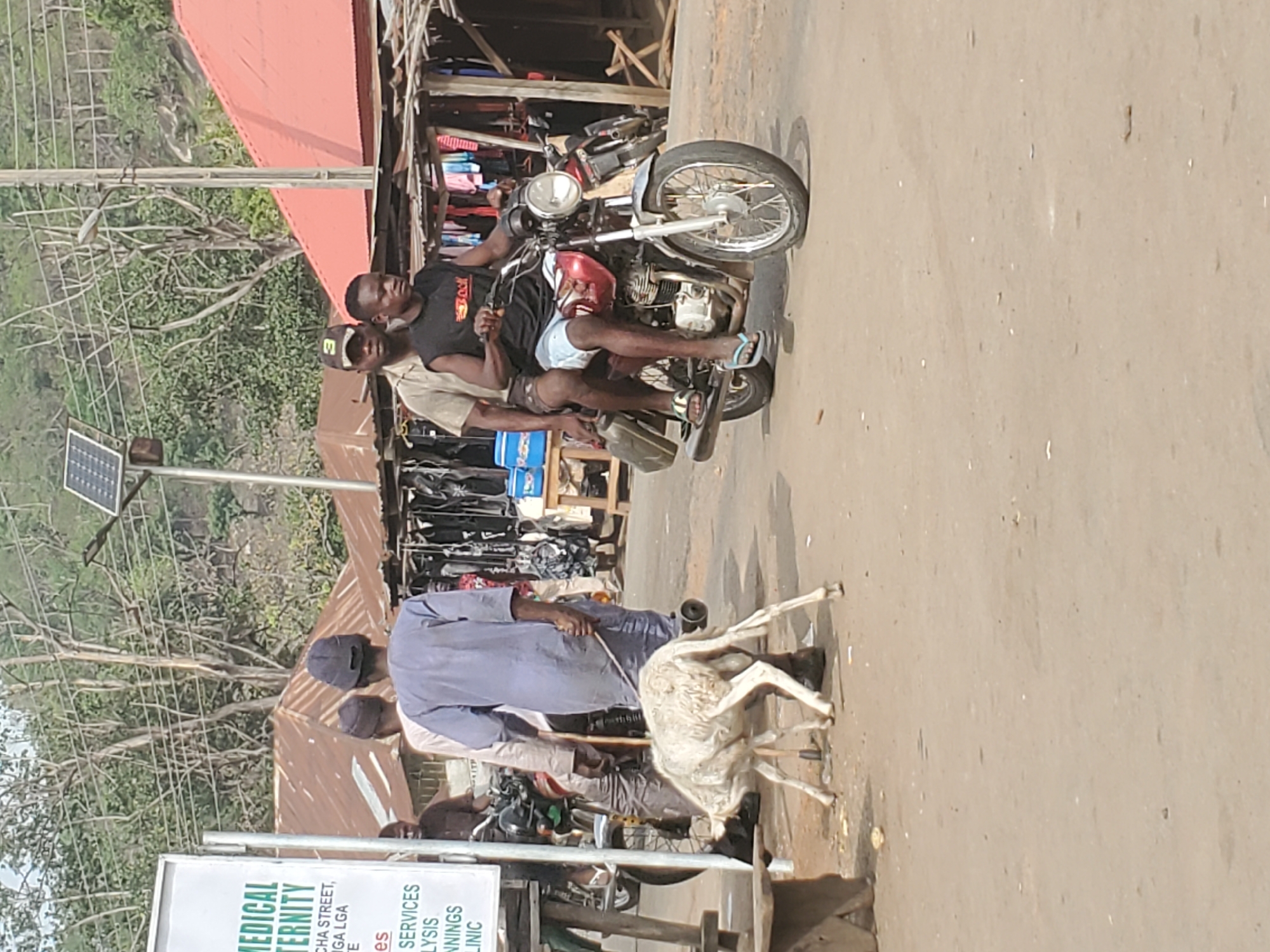 The image depicts a vibrant daytime street scene in what appears to be a market or semi-urban area, likely in Mararaba, Nigeria.
In the mid-ground, two men are on a red and black motorcycle. The driver, wearing a black t-shirt with a visible "YouTube" logo and light-colored shorts, is steering. A passenger in a beige top and dark cap sits behind him. The motorcycle is equipped with flip-flops on the footrests.
To the left, a man in a long, light blue garment is leading a white goat by a leash on the dusty ground. Another person is partially visible near him. In the background, behind the motorcycle and people, are several market stalls constructed with wood and corrugated iron roofs, displaying various goods. The stalls line what seems to be an unpaved road or open area.
Further in the background, green vegetation, trees, and hilly terrain are visible under a bright sky. A utility pole with a small solar panel attached is prominent on the left side of the image.
In the bottom left corner, a white signboard is partially visible. Clear text reads "MEDICAL MATERNITY," with smaller lines below including "CHA STREET," "IGA LGA," "SERVICES," "NNINGS," and "CLINIC," indicating a medical facility. The scene captures daily life with people, animals, and commerce coexisting in an active outdoor environment.