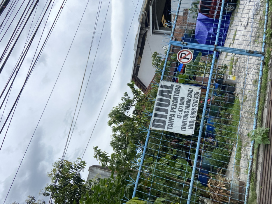 The image shows a blue metal fence in Margahayu, Indonesia, with a sign attached to it that reads "DIJUAL TANPA PERANTARA LT: 471 SHM (DIHITUNG TANAH SAJA) HUB: 0898 5710 335." There is a no parking sign visible through the fence. Green plants are growing on the outside of the fence and behind the fence. The sky is cloudy. The ground is gray and appears to be a concrete or brick patio. There are multiple power lines overhead.