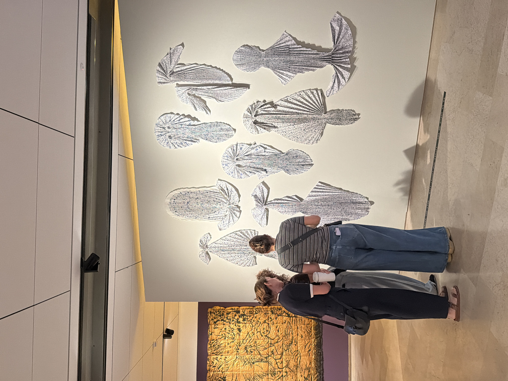 Two people stand in a room, likely an art gallery or museum, observing an installation of white, folded paper shapes resembling fish mounted on a wall. One individual wears a striped shirt and blue jeans, while the other wears a dark top, light grey pants and brown sandals. A large tapestry or wall hanging, predominantly gold and textured, is visible on the wall behind them. The room appears to be well-lit, with spotlights illuminating the artwork. The walls are white, and the floor has a light-colored, marbled or tiled appearance. The location is likely in the city of Tel-Aviv, Israel.