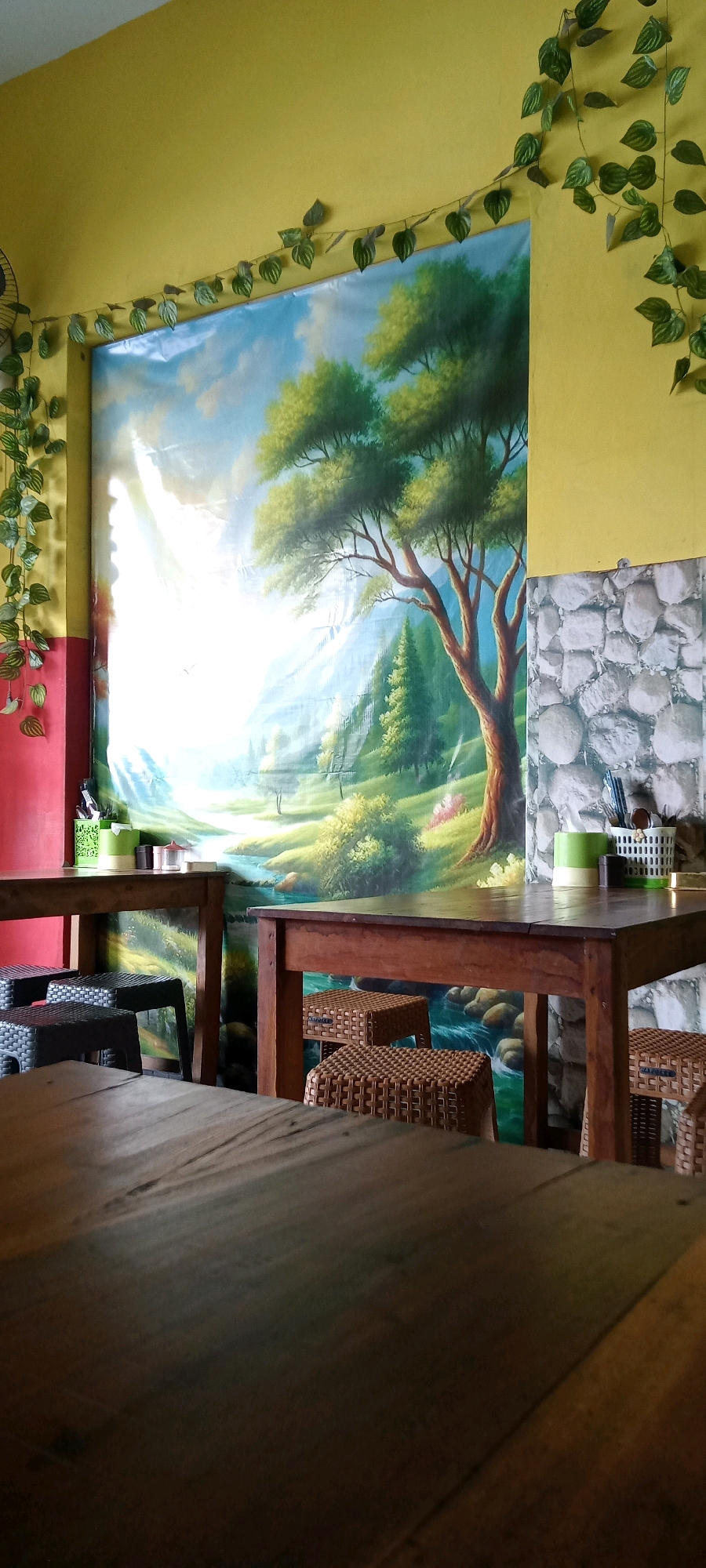The image depicts a dining area inside a building, likely a restaurant or cafe, with a large mural on the back wall. The mural features a scenic landscape with a large tree, mountains, and a river under a bright sky. The walls are painted yellow, with red on the left side. Artificial green vines hang from the ceiling and walls, adding a decorative touch.
In the foreground, there are several wooden tables and chairs. Two tables are visible: one on the left and one on the right. The table on the left has black plastic stools with a woven texture underneath it. The table on the right has brown woven stools. The table in the immediate foreground is wooden and appears to be a part of a larger table or another dining set.
On the table to the left, there are a few items: a green container with utensils, a small glass, and another small object. On the table to the right, there is a white basket with utensils and two green containers. The wall to the right of the mural has a stone-patterned wallpaper.
The lighting suggests an indoor environment, possibly with natural light coming from the left, illuminating the mural. The overall impression is a casual dining space decorated with a nature-themed mural.
Based on the provided information, this scene is located in Pondok, Indonesia.