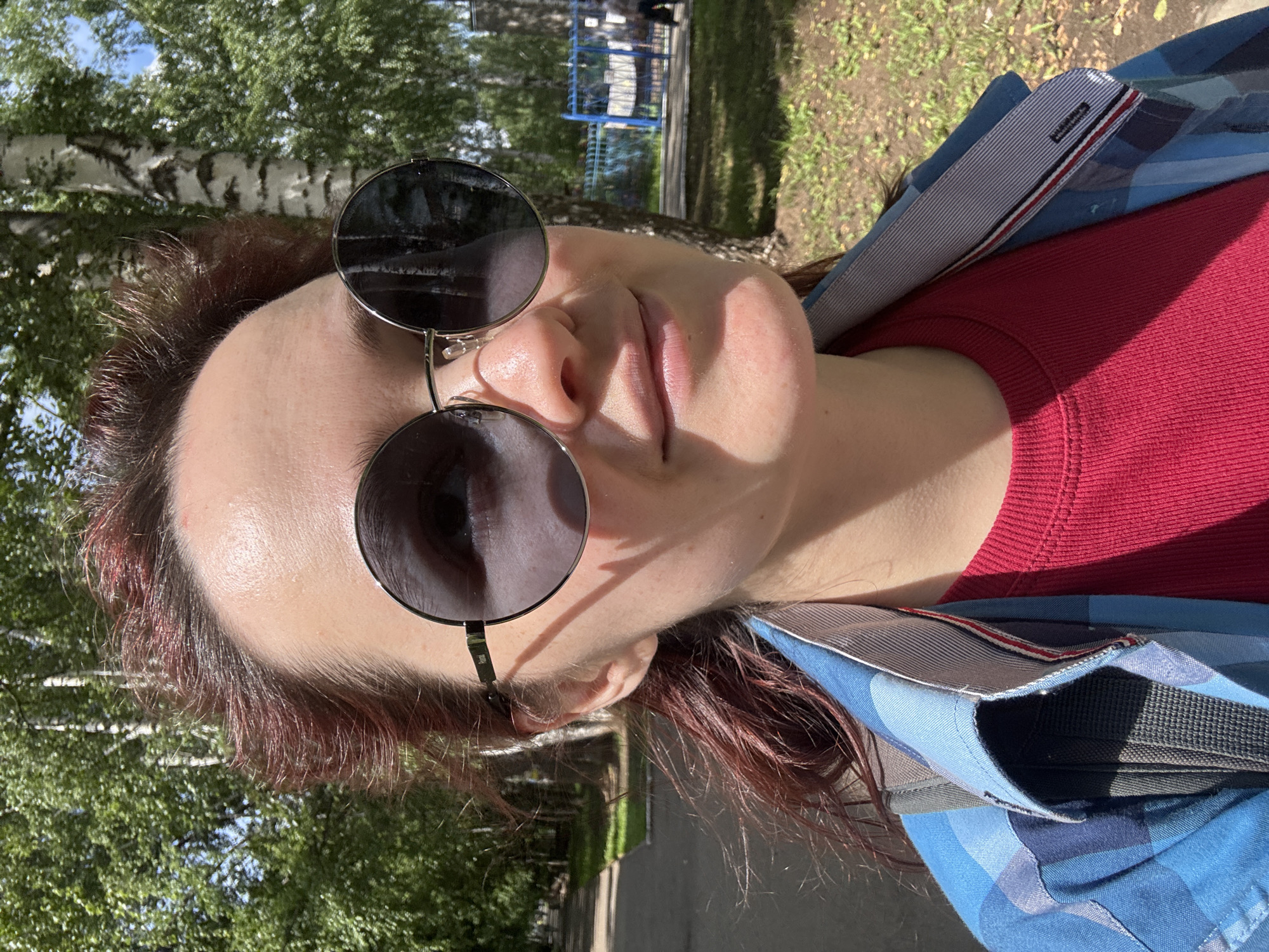 A young woman with auburn hair, wearing round sunglasses, a red t-shirt, and a blue plaid shirt, poses for a selfie outdoors in what appears to be a park or residential area in Izhevsk, Russia. The background features trees, a paved walkway, and a glimpse of a blue fence and buildings. The lighting suggests it is daytime, with sunlight casting shadows. The woman is looking slightly up and to the right, with a subtle smile.
