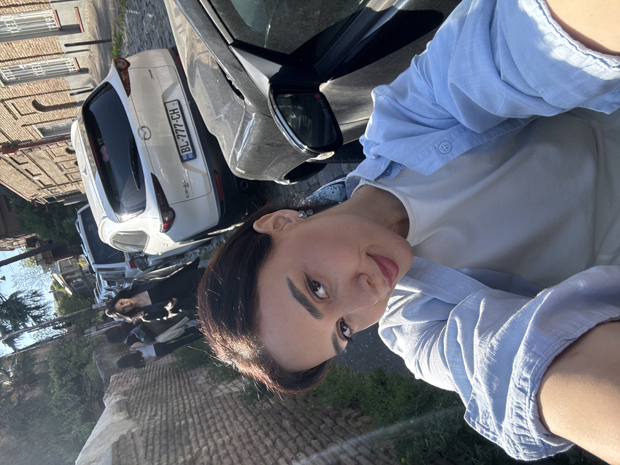 This outdoor selfie-style photograph, taken in Tbilisi, Georgia, captures a street scene under bright, sunny conditions.
In the foreground, a woman with dark hair and a slight smile looks towards the camera. She wears a light blue button-up shirt over a white top. A small, dark earring is visible on her left ear. Her face shows warm sunlight, suggesting morning or late afternoon.
Behind her, parked along the street, is a white Mazda CX-30 SUV. Its rear license plate clearly displays "BL-777-CH" and the "GE" country identifier. A small red sticker is faintly visible on the rear window of the Mazda. Partially visible to the right of the Mazda is a darker gray car. Further down the street, at least two other individuals can be seen in the mid-ground, one appearing to be another woman.
The background features brick or stone-clad building facades with windows, indicative of urban architecture. The ground in the foreground appears to be rough, possibly cobblestone or an uneven paved surface with some vegetation. Trees and plants line parts of the street, and more parked vehicles are visible further in the distance. The overall scene depicts a casual moment on a pleasant, sunny day in an urban environment.