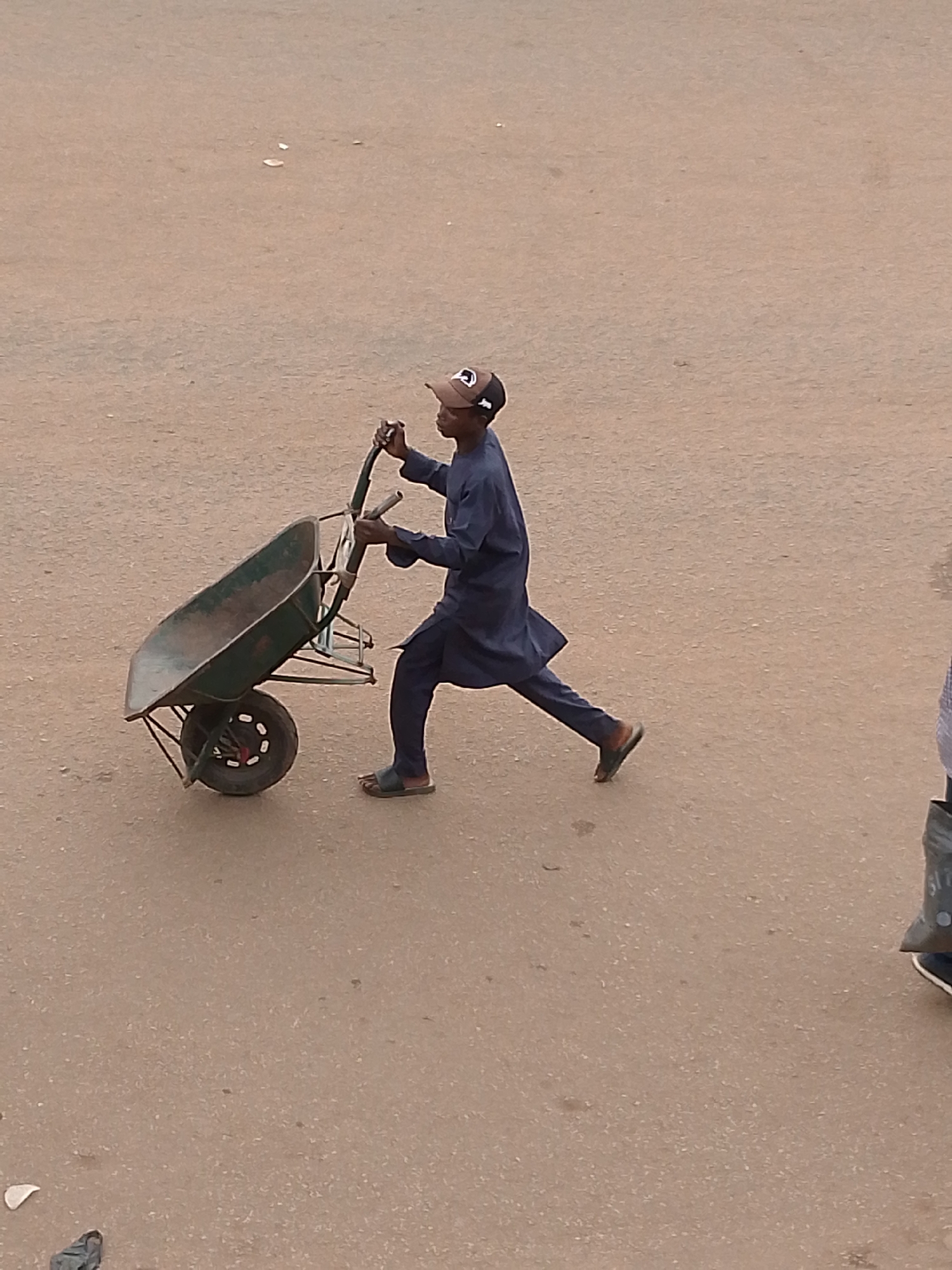 A young man is pushing an empty, green wheelbarrow on what appears to be a paved street in Bukuru, Nigeria. He is wearing a dark blue tunic and trousers, a baseball cap, and sandals. The pavement is cracked and stained. In the lower right corner, part of another person is visible. A discarded piece of white trash is visible in the lower left corner.