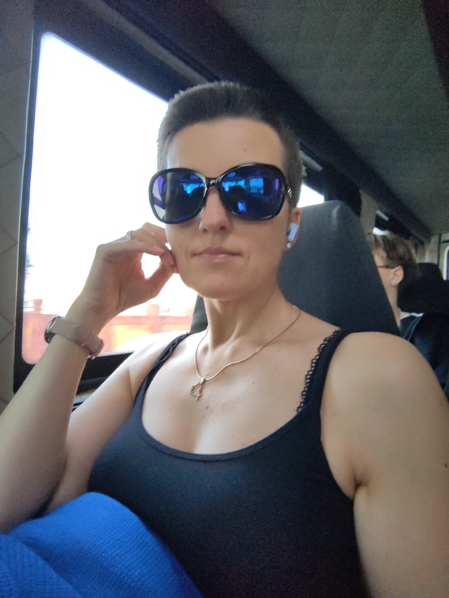 A woman with short dark hair, wearing sunglasses and a black tank top, is sitting in what appears to be a vehicle, possibly a bus or train, judging by the seats and window. She is wearing a silver necklace with a pendant and a smartwatch on her left wrist. Her right hand is resting on her chin, and she is looking towards the camera. The environment outside the window appears to be urban, with a blurred view of buildings. There is another person visible in the background, seated behind her. The lighting suggests it is daytime, possibly late afternoon. The location is identified as Minsk, Belarus.