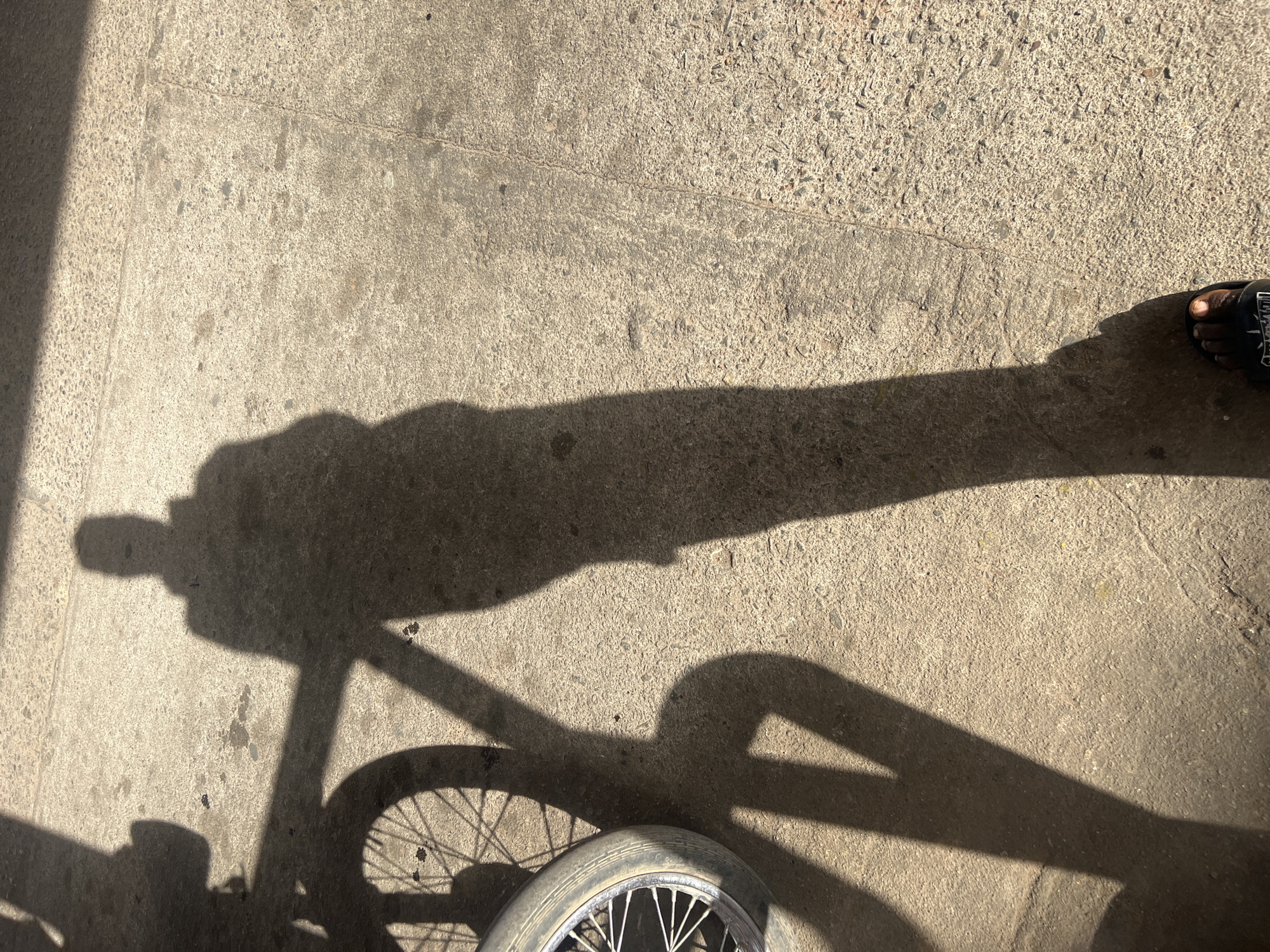 The image captures a ground-level view of a textured concrete surface, likely a pavement or road, illuminated by strong sunlight. Dominant in the frame are long, dark shadows cast across the concrete.
**Subjects:** The primary subjects are the shadows of a person and a bicycle. The person's shadow is long and stretches from the left side of the frame towards the right, suggesting a low sun angle. Distinct features of the person's shadow include what appears to be an arm and head. Below this, the shadow of a bicycle is clearly visible, showing parts of its frame, handlebars, and a front wheel with visible spokes. In the bottom right corner, a human foot, wearing a dark sandal, is visible on the sunlit concrete, casting its own small shadow.
**Activity or event taking place:** The scene depicts a person either standing next to or riding a bicycle, looking down at the ground. The long shadows suggest movement or presence on a sunny day.
**Notable details:** The long, defined shadows indicate that the photograph was taken either early in the morning or late in the afternoon when the sun is low in the sky. The bright concrete surface and sharp shadows suggest clear, sunny weather. The concrete itself has a rough, aggregate texture with some minor cracks and dirt stains. The sandal worn by the foot is dark with a white design or logo on the strap, which appears to be text, possibly "KAPPA", though it's partially obscured and not perfectly clear. Given the location context of Jos, Nigeria, this is a common urban or suburban ground setting in a sunny climate.
**Visible text:** The word "KAPPA" appears to be visible on the strap of the sandal, though it is slightly blurry and partially obscured.