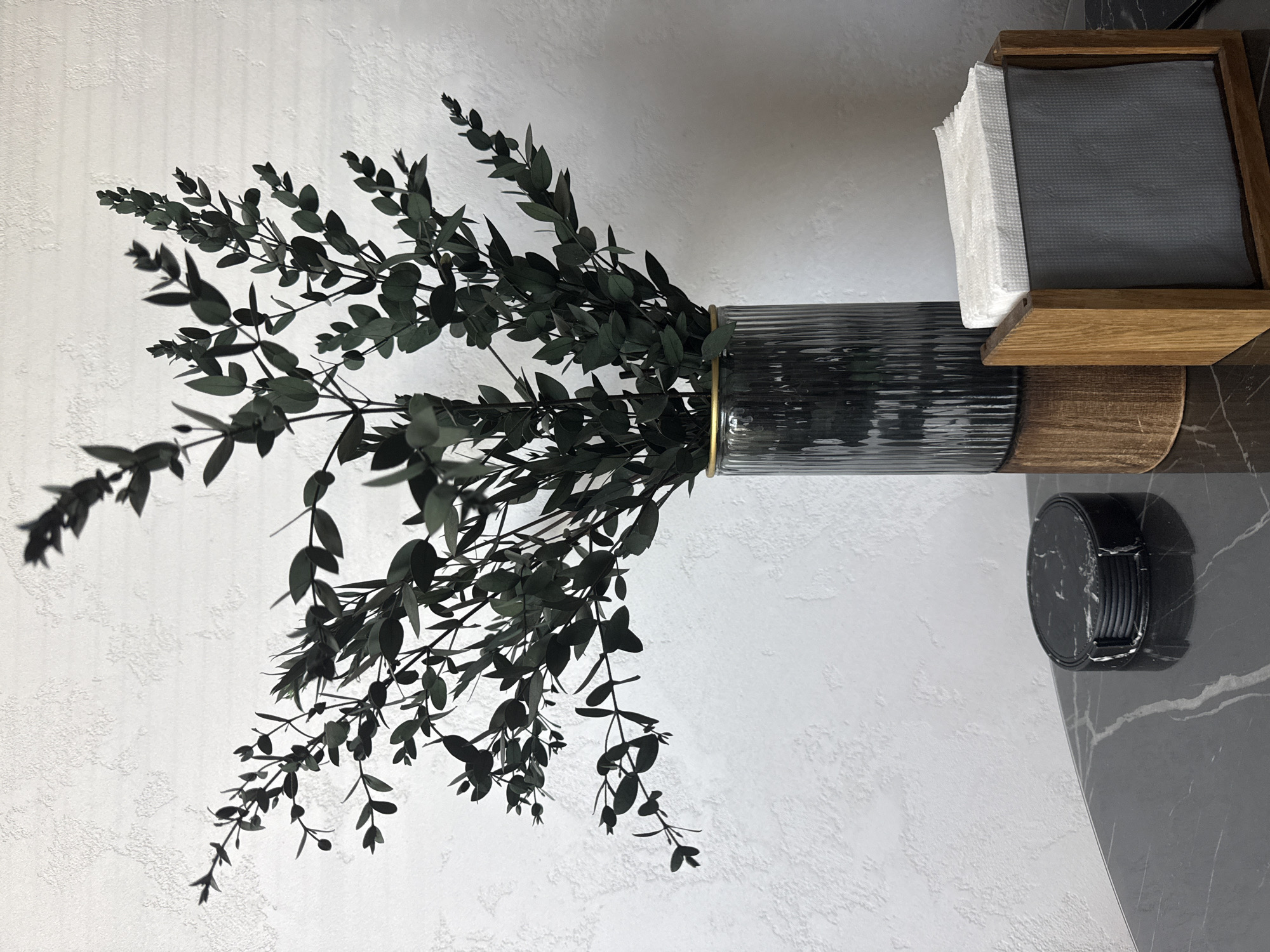 In an indoor setting, a dark green, leafy branch arrangement is displayed in a fluted glass vase with a gold rim. The vase rests on a wooden pedestal. To the left of the vase, on a black marble surface, is a stack of round, black marble coasters. To the right of the vase is a wooden napkin holder filled with white napkins. The background is a textured white wall. The scene is devoid of people and activity. The photograph appears to be taken in Velikiye Luki, Russia.