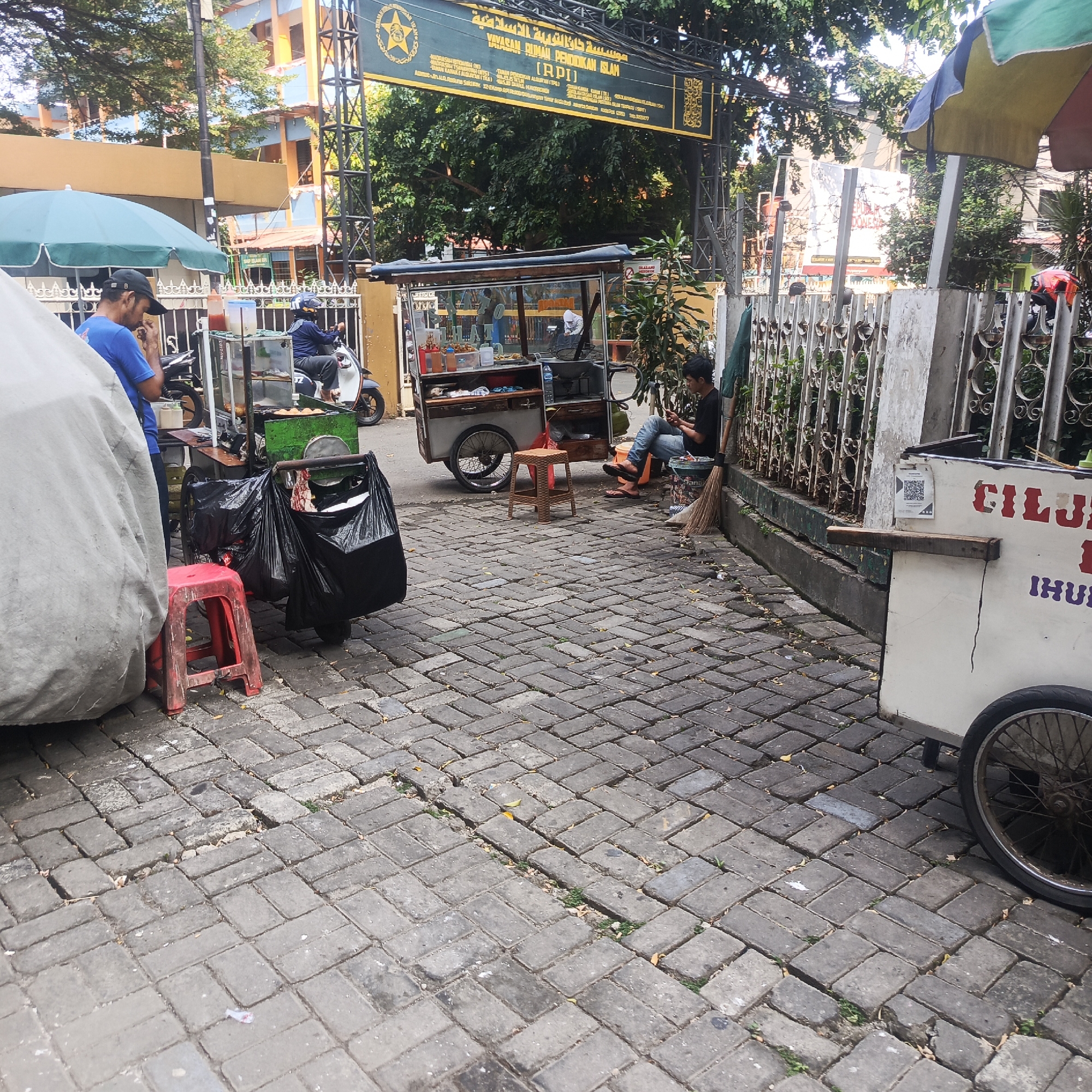 The image captures a street scene in South Jakarta, Indonesia, likely in an area with food vendors. In the foreground, a white cart with a bicycle wheel and red lettering is visible. To the left, a large, grey tarp covers an object, with a red plastic stool placed in front of it. Next to the tarp, a man in a blue shirt and black cap is partially visible, appearing to eat. A food cart with a glass display and various items inside is situated in the middle of the scene. To the right of the food cart, a young man in a black shirt and jeans is seated on the ground, looking at his phone, with a broom leaning against a metal fence behind him. A motorcycle is parked near the food cart, and another motorcycle is visible further back. The ground is paved with interlocking grey bricks. In the background, a building with a yellow and blue facade stands behind a metal gate, above which a green banner with Arabic and Indonesian text is displayed. Trees and foliage are visible behind the banner and gate. The overall atmosphere suggests a busy, informal commercial area within the city.