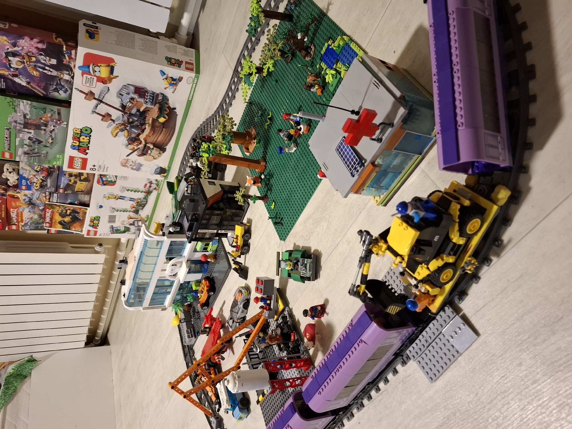 This image shows a collection of Lego models arranged on a light-colored wooden floor, likely indoors. The models depict a city scene with various buildings, vehicles, and figures. In the foreground, a purple Lego train with a yellow construction vehicle attached to it is on a track. Behind the train, there is a modern-looking Lego building with solar panels and a red cross symbol on its roof, resembling a hospital or clinic. To the left of the train is another Lego structure with an orange crane. Further back, a street scene unfolds with a white and blue Lego building, a car, and more construction vehicles. In the background, several Lego boxes are stacked, including those for "Lego Super Mario" and "Lego Minecraft." A white radiator is visible along the wall in the background. The overall arrangement suggests a playful and imaginative setup. There is no indication of the specific location being Tyumen, Russia.