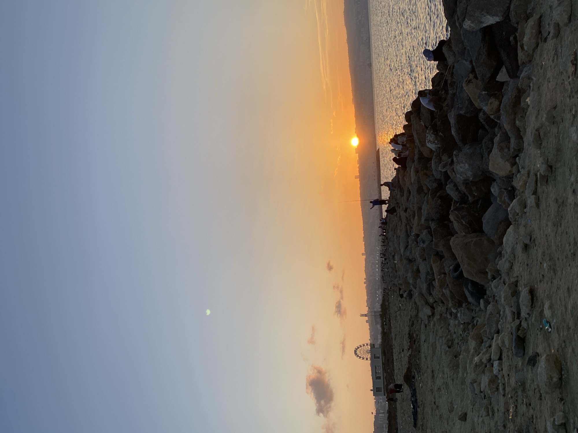 The image depicts a sunset over the coast of Mohammadia, Algeria. A rocky embankment extends into the sea, where several individuals are visible. Some are engaged in fishing, while others are seated and observing the scenery. The sun is low on the horizon, casting a warm orange glow across the sky and water. In the background, a Ferris wheel and city buildings are silhouetted against the sky. The foreground consists of a sandy beach littered with rocks and debris. The overall atmosphere is peaceful and serene, with a sense of community as people gather to enjoy the evening by the sea.