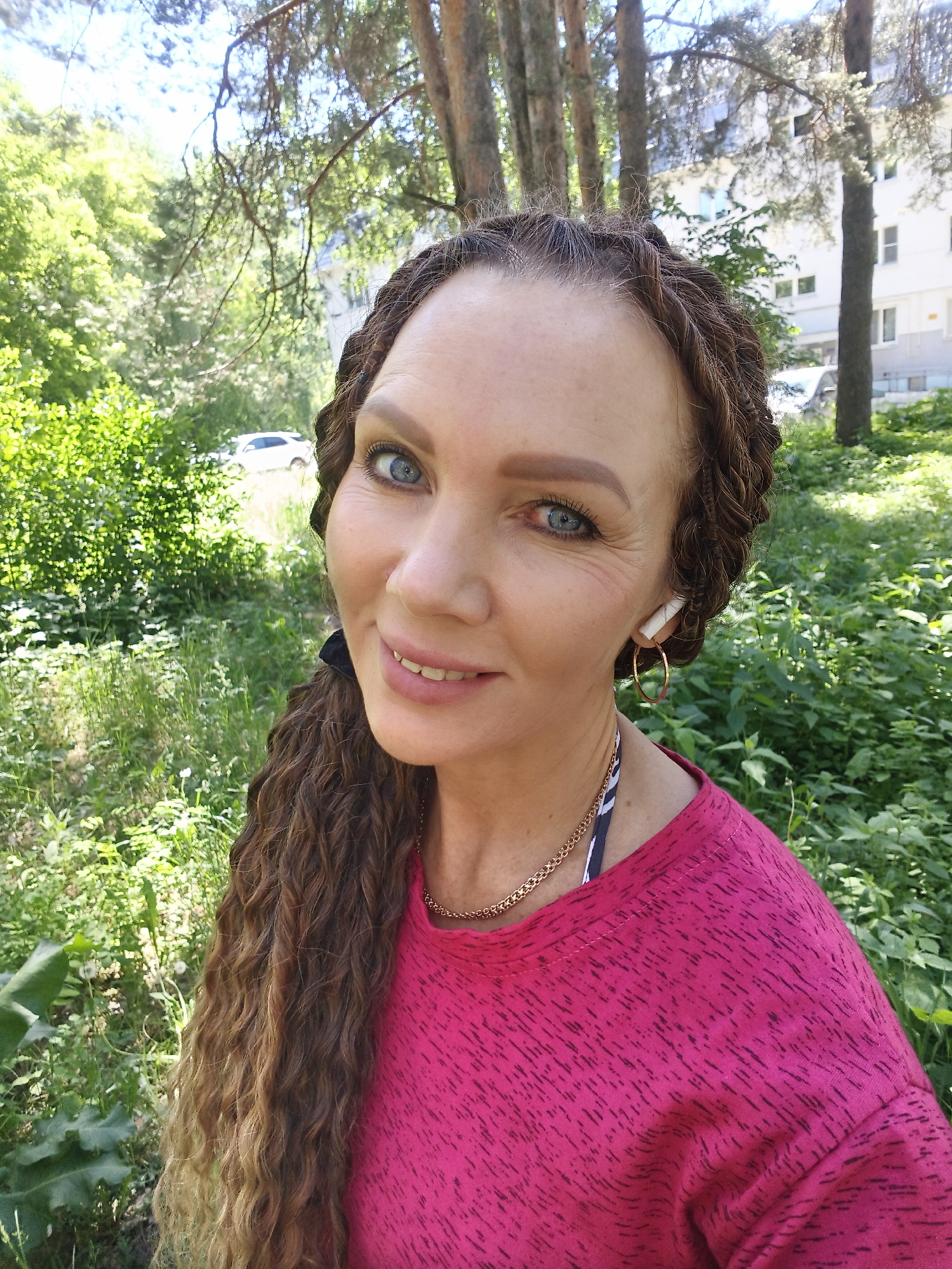 A woman with long, braided brown hair and blue eyes is taking a selfie outdoors. She is wearing a pink shirt, gold hoop earrings, and a white earbud. She is smiling at the camera. The background is a lush green area with trees and buildings. It is daytime and the sun is shining.