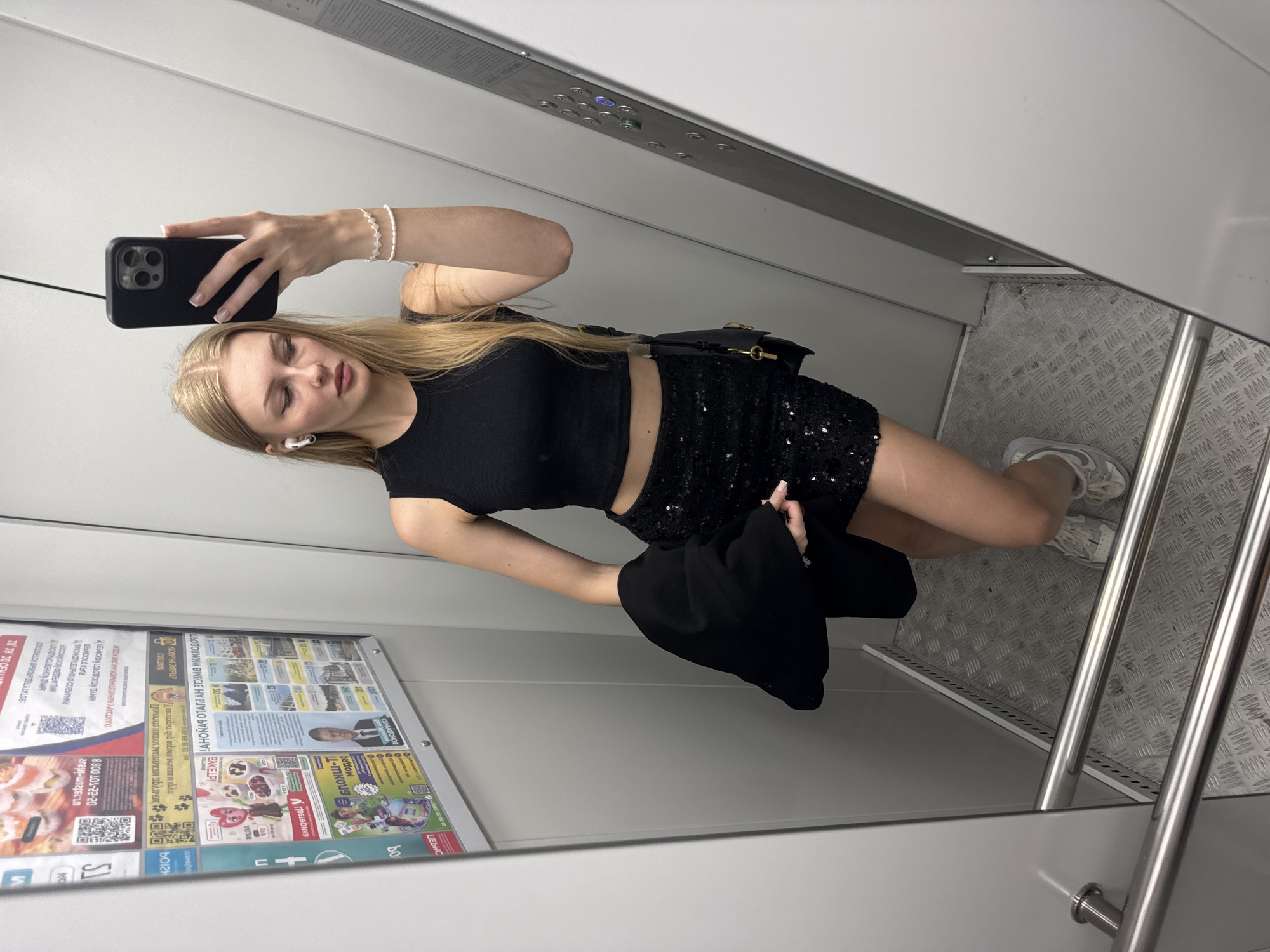 A young woman with blonde hair, wearing a black crop top, a sparkly black skirt, white sneakers, and holding a black jacket, stands in an elevator and takes a selfie. She has on white earbuds and a beaded bracelet. The elevator is utilitarian with grey walls and a diamond plate metal floor. There are posters on the left wall, some with QR codes and text in Russian, suggesting the location is in Russia. One poster has text that reads "Perm".