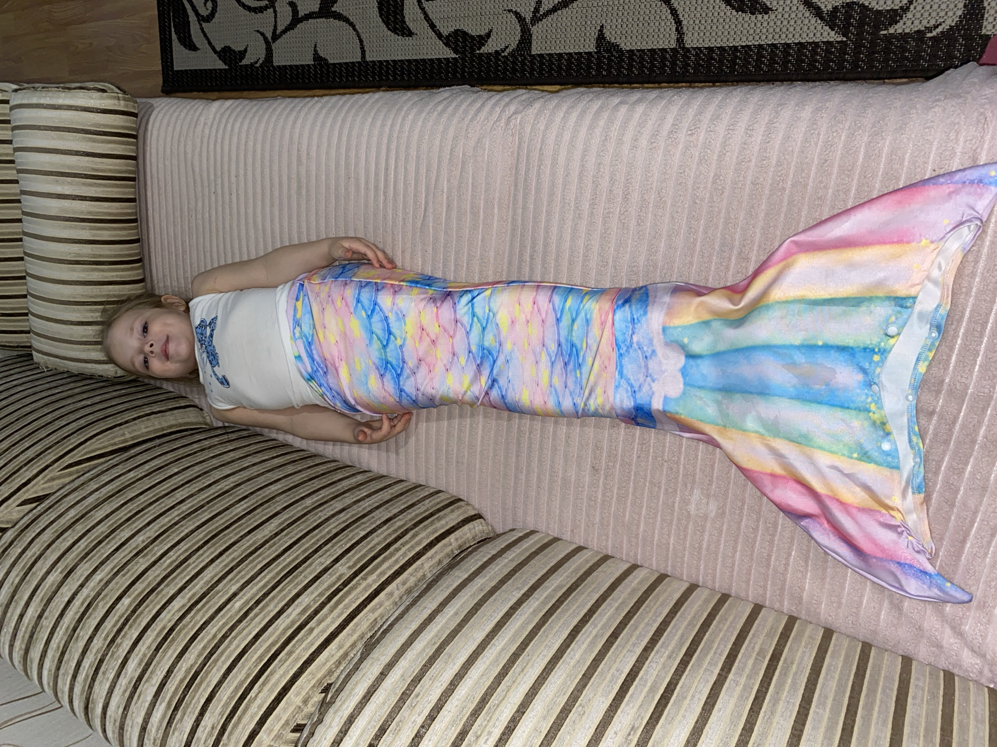 A young child is lying on a light-colored sofa in an indoor setting, likely a home in Votkinsk, Russia. The child, with light hair, is wearing a white t-shirt with a blue pattern on the front, and a vibrant, multi-colored mermaid tail costume. The mermaid tail features a scale pattern in shades of pink, blue, and yellow near the top, transitioning into horizontal rainbow-like stripes towards the fin, which is a blend of pink, blue, and purple.
The child is positioned with her head on a light pink/beige ribbed section of the sofa, and her body extends across the striped seating area, which has beige and brown stripes. She is smiling and looking directly at the camera, conveying a happy and relaxed mood. Her arms are bent, resting on her stomach area.
In the background, behind the sofa, a dark rug or mat with a patterned design is partially visible. The general lighting suggests it is daytime. No discernible text is visible in the image.