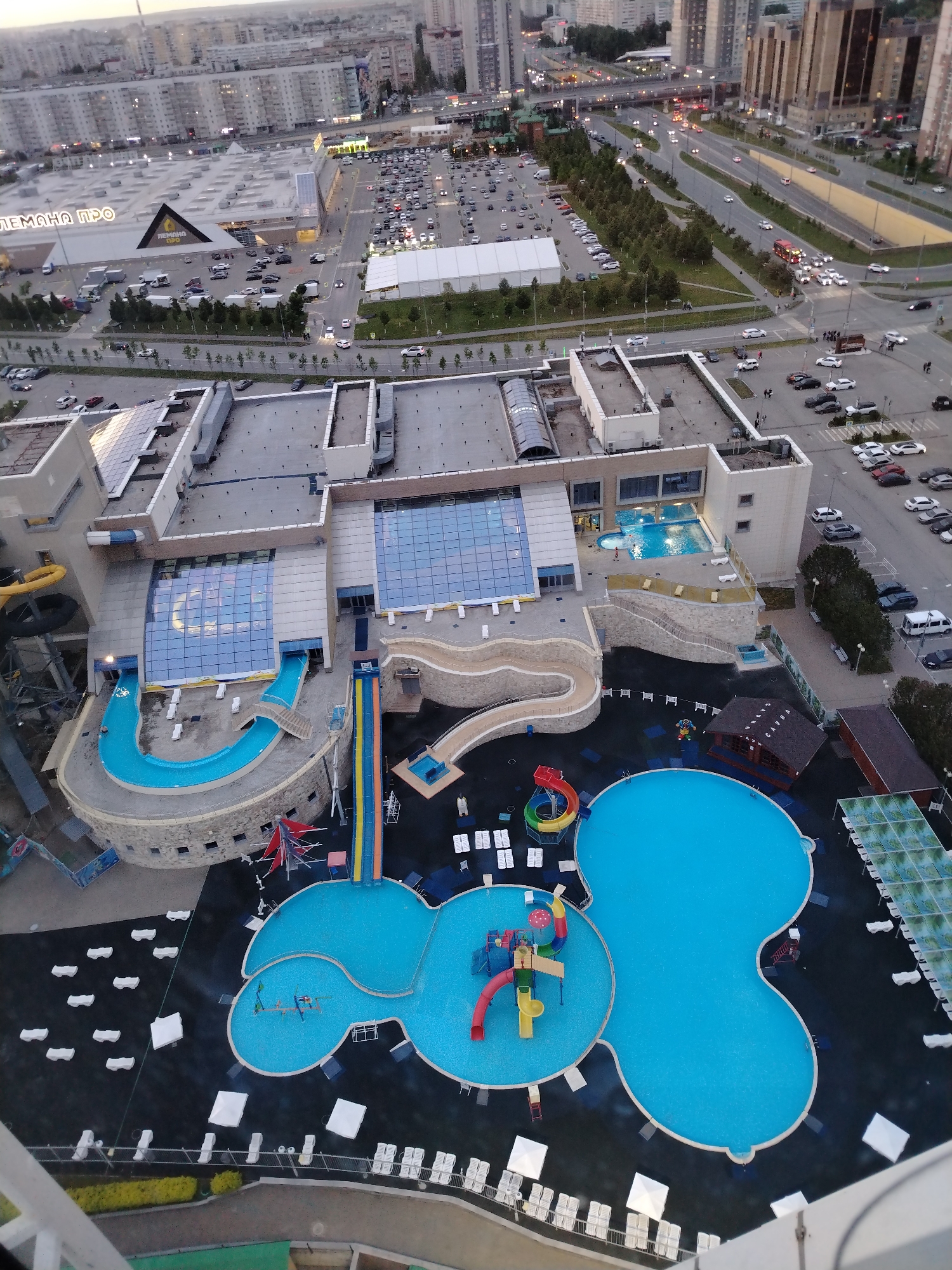 An aerial view shows the Riviera Aquapark in Kazan, Russia. The waterpark features numerous outdoor and indoor pools, water slides, and leisure areas. People are visible swimming in some of the pools. In the background are apartment buildings, a shopping mall, and a highway with vehicular traffic. The overall image conveys a sense of a recreational facility in an urban environment.