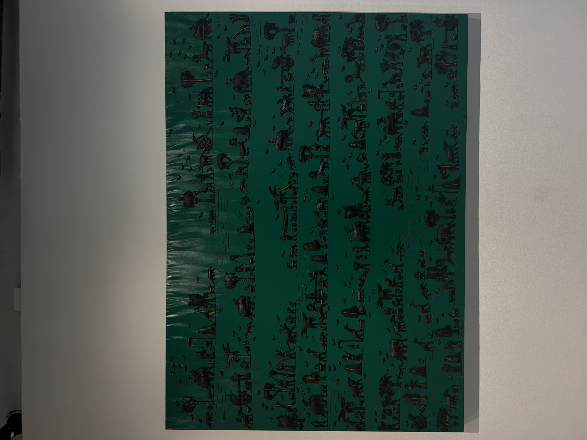 This is a photograph of a large, dark green, textured artwork mounted on a white wall. The artwork appears to be a detailed, frieze-like composition filled with numerous small, silhouetted figures of animals and people. The figures are arranged in horizontal rows, creating a sense of narrative or a dense, populated landscape. The style is illustrative and somewhat cartoonish, with many of the animal figures resembling stylized elephants, giraffes, lions, and other creatures. Some human figures are depicted interacting with the animals or engaged in various activities. The overall impression is of a busy, fantastical scene, possibly depicting a wildlife sanctuary, a fantastical jungle, or a narrative across different strata. The lighting suggests an indoor setting, likely a gallery or museum space, given the white wall and the way the artwork is presented. The background wall is smooth and clean, with no other objects visible apart from a small white rectangular object near the top edge of the image. The bottom of the artwork meets the top of a white, slightly textured wall which occupies the lower half of the frame. The top portion of the image shows a hint of a dark, possibly black, object in the upper left corner, and a white object near the center, suggesting the presence of lighting fixtures or ceiling elements. The texture of the artwork itself seems to be composed of small, vertical ridges, giving it a ribbed or corrugated appearance. The detailed nature of the artwork suggests it could be a relief carving, a highly detailed print, or a mixed-media piece. There is no visible text or signage in the image that would indicate the specific location.