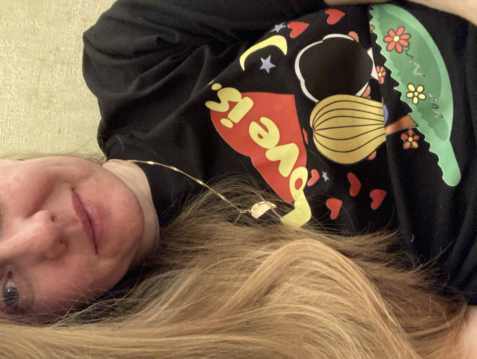 A woman lies on her back, possibly on a bed, indoors. She wears a black t-shirt with a colorful graphic depicting a person with a yellow ponytail next to a green leaf with flowers. The shirt has the phrase "Love is" on it, written in red and yellow. The background appears to be a textured wall. She wears a gold necklace with a small pendant. Her hair is long and blonde. The image was not taken in the city of Chaikovsky, Russia.