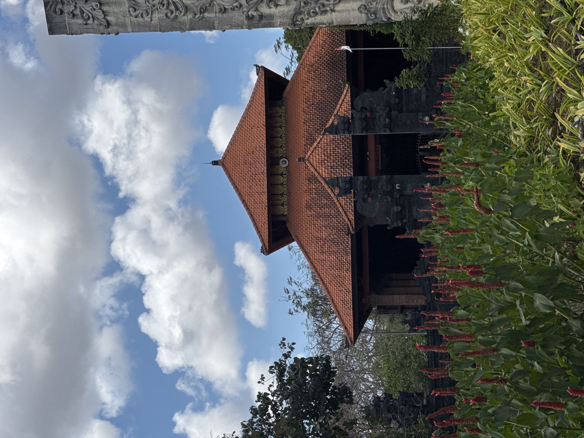 In Kuta Selatan, Indonesia, a vibrant outdoor scene unfolds under a partly cloudy blue sky. In the foreground, a lush bed of red-tipped green plants bursts with life, their tall stalks reaching upwards. Behind them, a traditional Balinese temple with a striking orange tiled roof takes center stage. The temple's dark, intricately carved stone entrance is flanked by ornamental statues, suggesting a place of cultural or religious significance. To the right, a tall, ornate stone pillar rises, its surface detailed with intricate carvings, partially obscuring the temple's facade. A small Indonesian flag flutters gently on a pole near the temple entrance, adding a touch of national pride to the scene. In the background, dense green foliage and trees frame the temple, indicating a natural, verdant setting.
