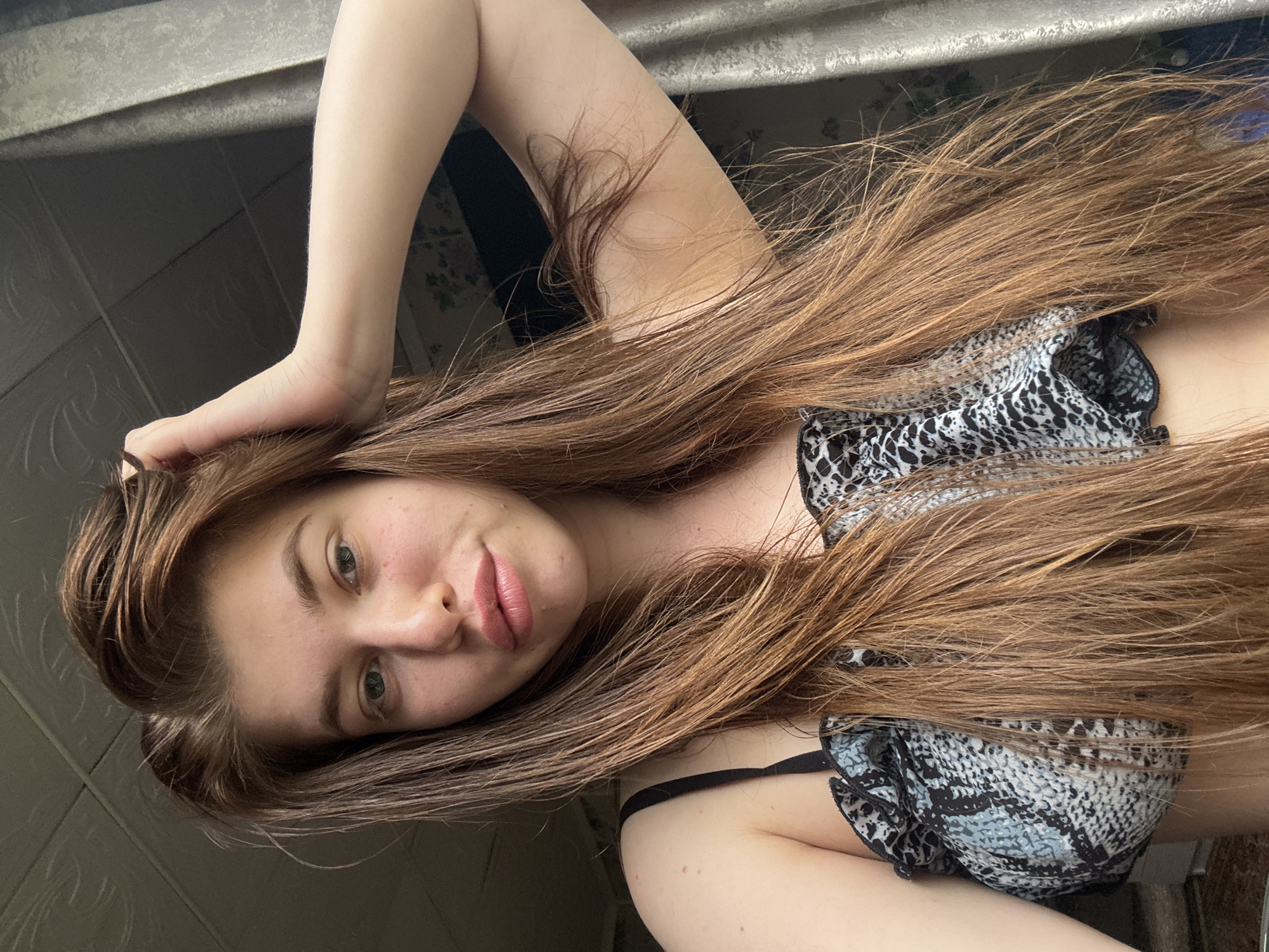 A woman is reclining, possibly indoors. She has long, brown hair and is wearing a patterned top or bikini. One arm is raised with her hand near her hair, and the other arm is visible. The background appears to be a room interior. There is no evidence that this image was taken in the city of Meshchery, Russia.