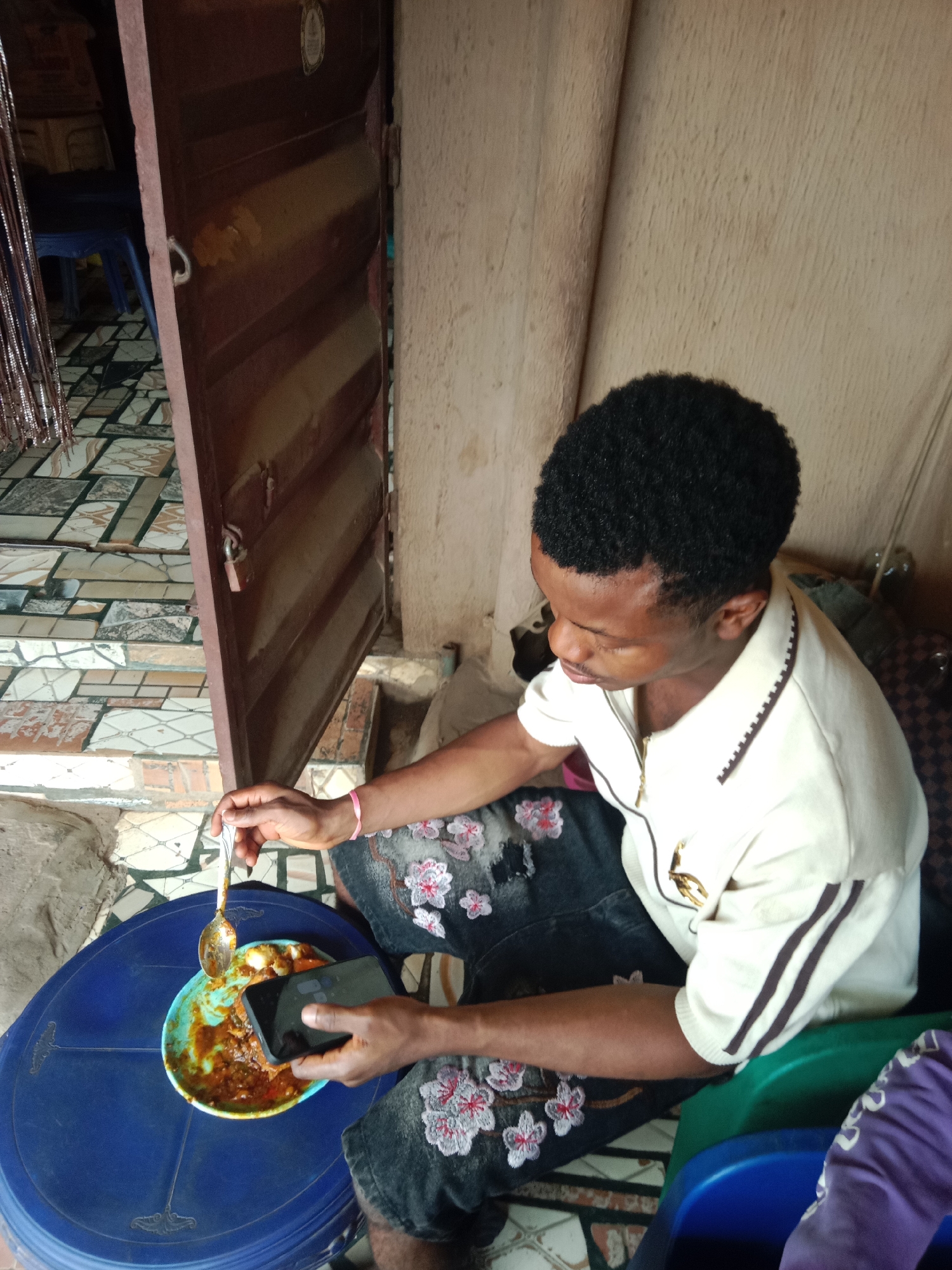 In Onitsha, Nigeria, a young adult male with short dark hair is seated, holding a spoon with food over a light blue plate that rests on a dark blue plastic stool. He wears a cream polo shirt with brown stripes and dark denim shorts featuring white and pink floral embroidery, with a pink wristband on his right wrist. His left hand holds a black smartphone, and he looks downwards towards the food/phone. The background consists of a light brown textured wall to the right and a partially open dark brown metal door to the left, revealing a patterned tiled floor, a blue plastic chair, and a silver fringe curtain.