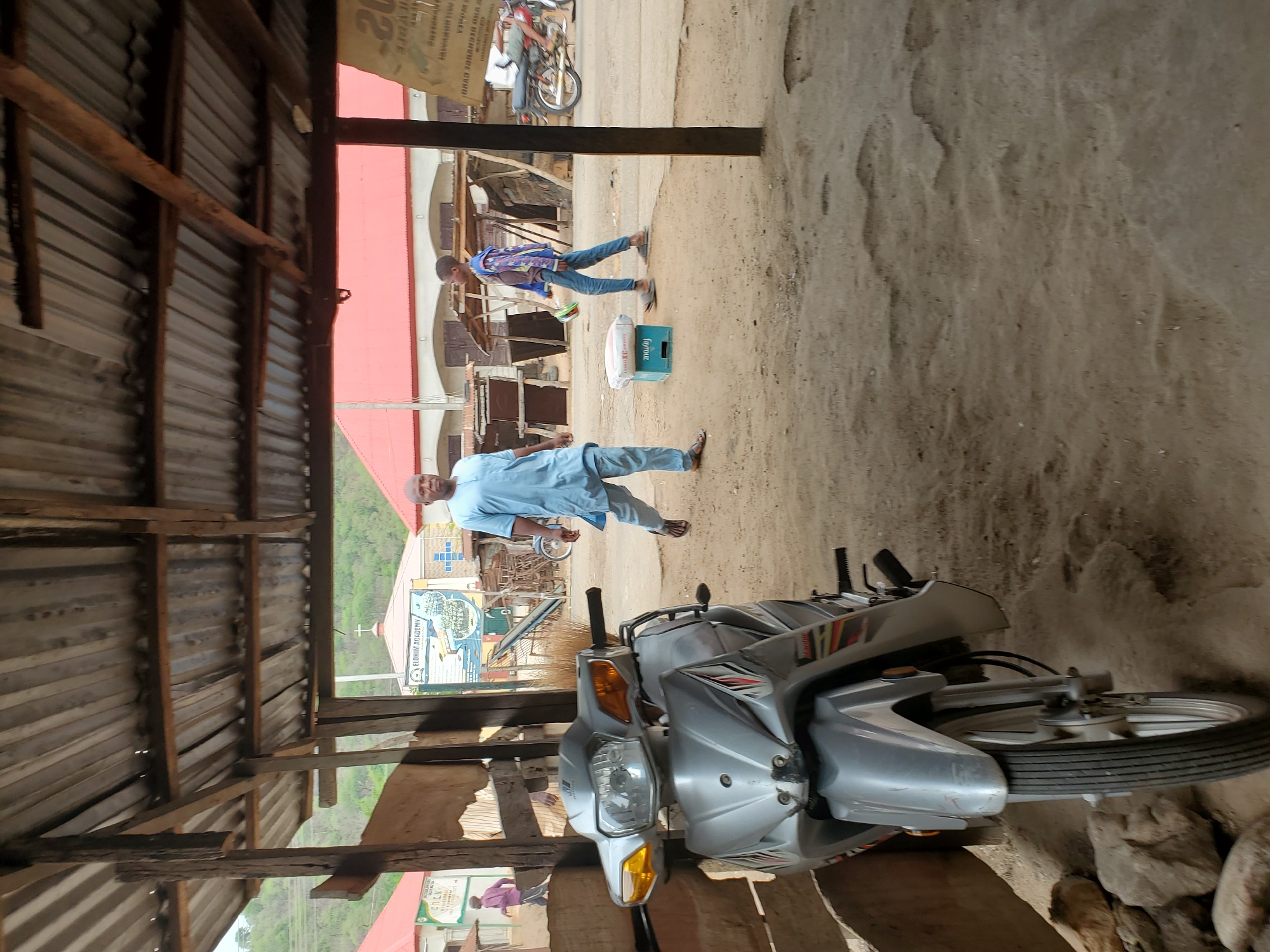The scene is an outdoor shot in Mararaba, Nigeria, taken from beneath a corrugated metal roof structure with exposed wooden supports. A silver motorcycle dominates the foreground. An adult male wearing a light blue ensemble walks across the open space. Further back, another individual in a blue patterned shirt and jeans is seen walking past a blue crate and a white sack. In the background, the "ELOHIM ACADEMY" sign is visible.