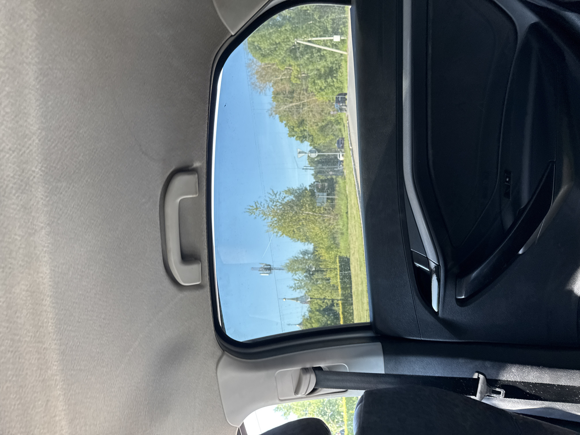 The image shows the interior of a vehicle looking out of a rear window. The interior consists of a grey headliner, a grey grab handle, a black seatbelt, and a black car door with a silver door handle. Outside the window, the sky is clear and blue. A row of trees is visible in the middle ground, with a road and more trees in the background. A cell tower can be seen in the distance. The scene appears to be located in Chaikovsky, Russia, based on the distinct architecture of the church spires visible amongst the trees.