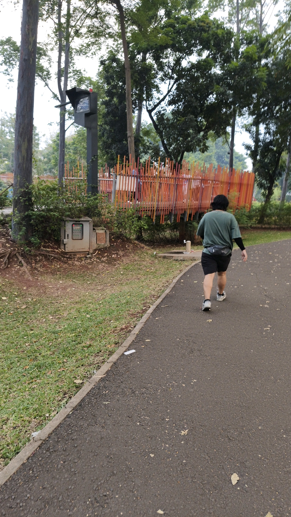 The image depicts a paved pathway in a park setting, likely in South Jakarta, Indonesia, with lush greenery and trees surrounding it. A person, seen from behind, is walking away from the camera on the pathway. The individual is wearing a dark green t-shirt, black shorts, and athletic shoes. They also have a black fanny pack around their waist and a black arm sleeve on their right arm. To the left of the pathway, there's a grassy area with fallen leaves and tree roots visible. Further left, a metal utility box with a keypad is situated near the base of a tree. In the background, there's an orange and yellow striped fence structure, possibly part of a playground or park amenity, with more trees and foliage behind it. A tall, dark pole with a camera or sensor is also visible among the trees. The overall environment suggests an outdoor recreational space.