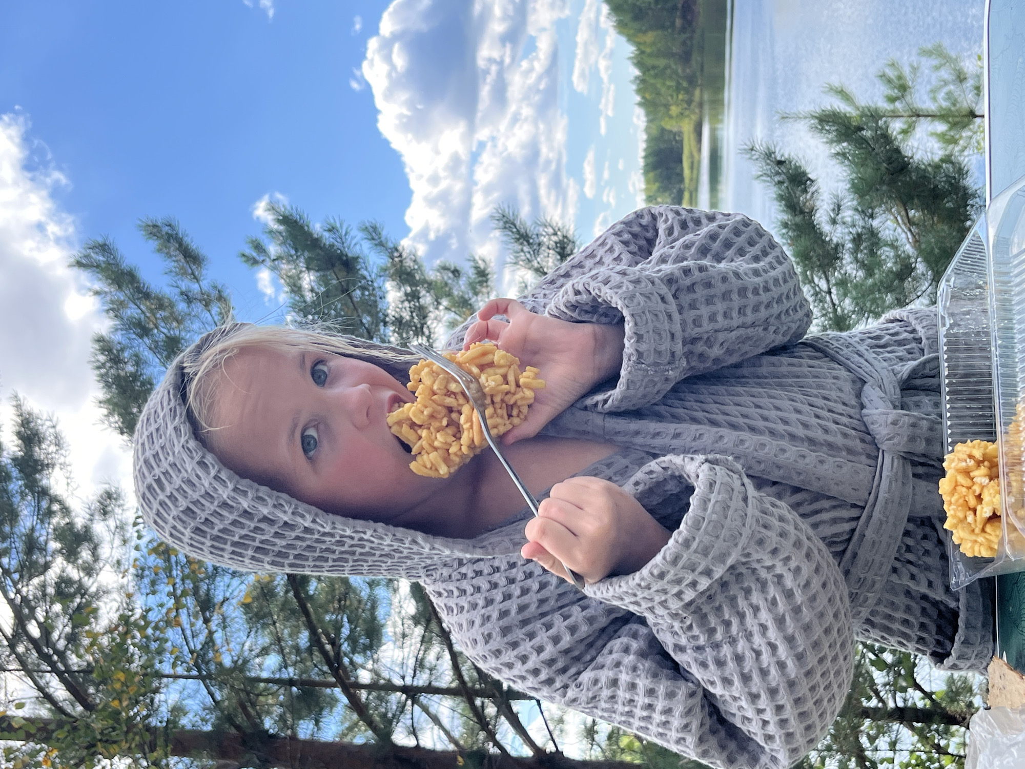A young blonde girl wearing a grey waffle-knit hooded robe is eating a sweet, fried dough pastry from a fork. She is outdoors, with a body of water and trees in the background. The sky is blue with scattered white clouds. The location is Novyi, Russia.