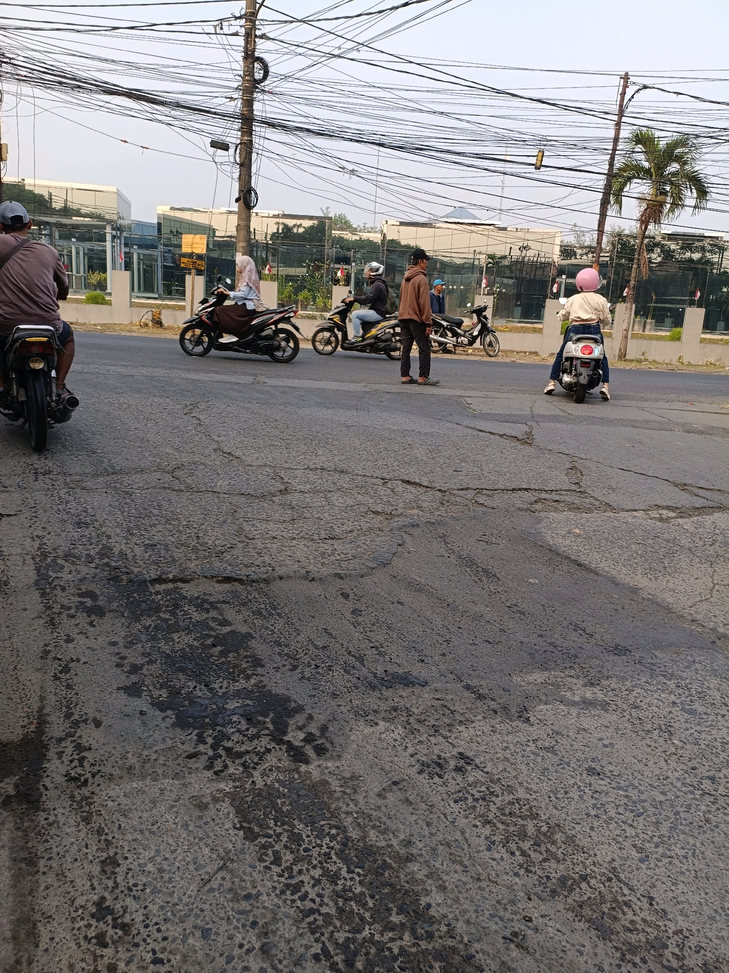 The image depicts a street scene in Tangerang, Indonesia, with multiple motorcycles and riders. In the foreground, a motorcycle rider wearing a gray shirt and a dark cap is partially visible from the rear. To the left, another motorcycle with a rider wearing a light brown jacket and dark pants stands near a parked motorcycle. Further ahead, a motorcycle rider in a light-colored jacket and dark pants with a pink helmet is visible from the rear, as is a motorcycle rider in a light blue shirt and brown skirt. The background shows modern buildings with reflective glass facades, a palm tree, and an overhead network of electrical wires. The road surface is asphalt, showing signs of wear and tear with patches of dark stains and cracks. The overall scene suggests a typical urban environment with bustling traffic.