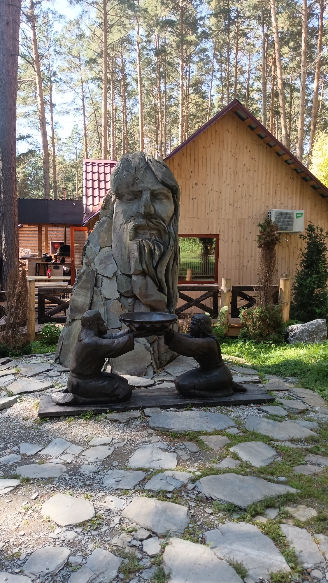 This is a medium shot, outdoor, daytime photograph of a sculpture garden. In the foreground, two dark brown, wooden statues of kneeling figures hold a shallow bowl between them. Behind the statues, a large, grey stone sculpture of a man's head with a beard is positioned. To the right of the stone sculpture is a wooden building with a gabled roof and a light brown exterior. An outdoor air conditioning unit is mounted on the wall of the building. A wooden fence with cross braces is visible in front of the building. The ground is paved with irregular, grey flagstones and gravel, with patches of green grass growing between them. In the background, tall, thin pine trees form a dense forest. The sky is a clear, light blue.