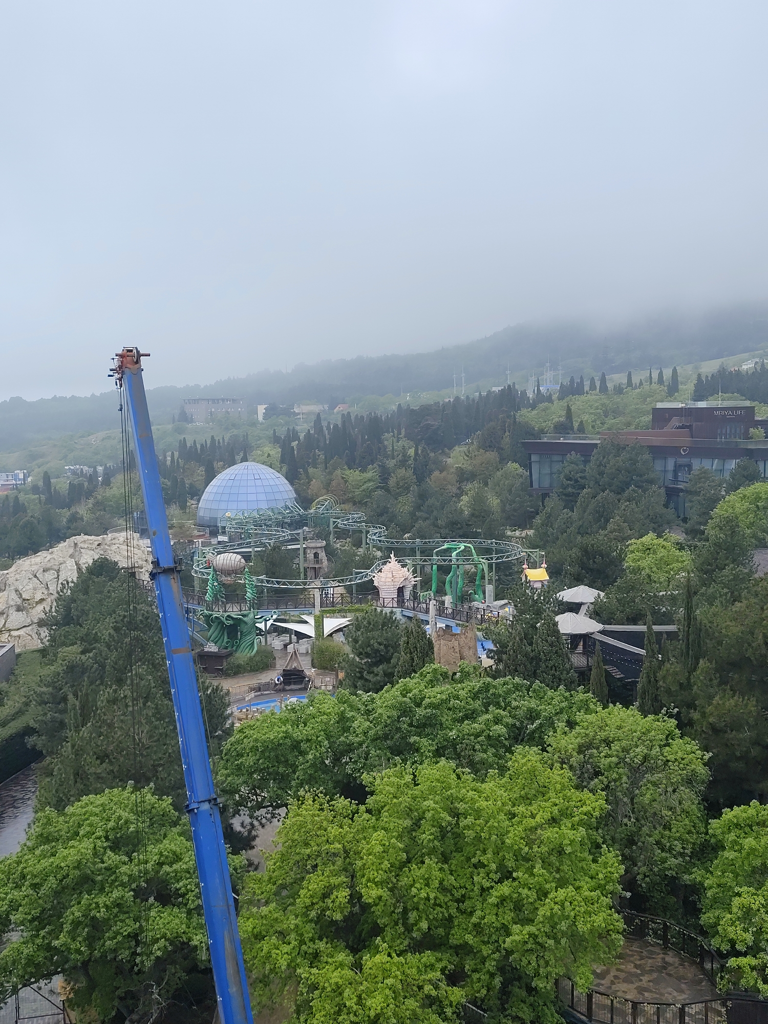 Captured from an elevated perspective on an overcast day in the city of Симеизский поселковый совет, Ukraine, the image showcases a blend of natural and constructed elements.
A large, blue crane takes up a significant portion of the left foreground, its extended boom intruding into the scene. Below the crane, verdant treetops form a dense canopy, partially obscuring features beneath.
In the center of the frame, a whimsical amusement park appears, complete with a bright blue geodesic dome and an elaborate roller coaster winding around a whimsical castle-like structure. Other rides and attractions are visible, though somewhat veiled by the abundant greenery.
In the background, a mountainous landscape rises. Buildings, including one labeled "MRIYA LIFE," dot the landscape. The overcast sky casts a muted light over the entire scene, adding to the atmosphere.