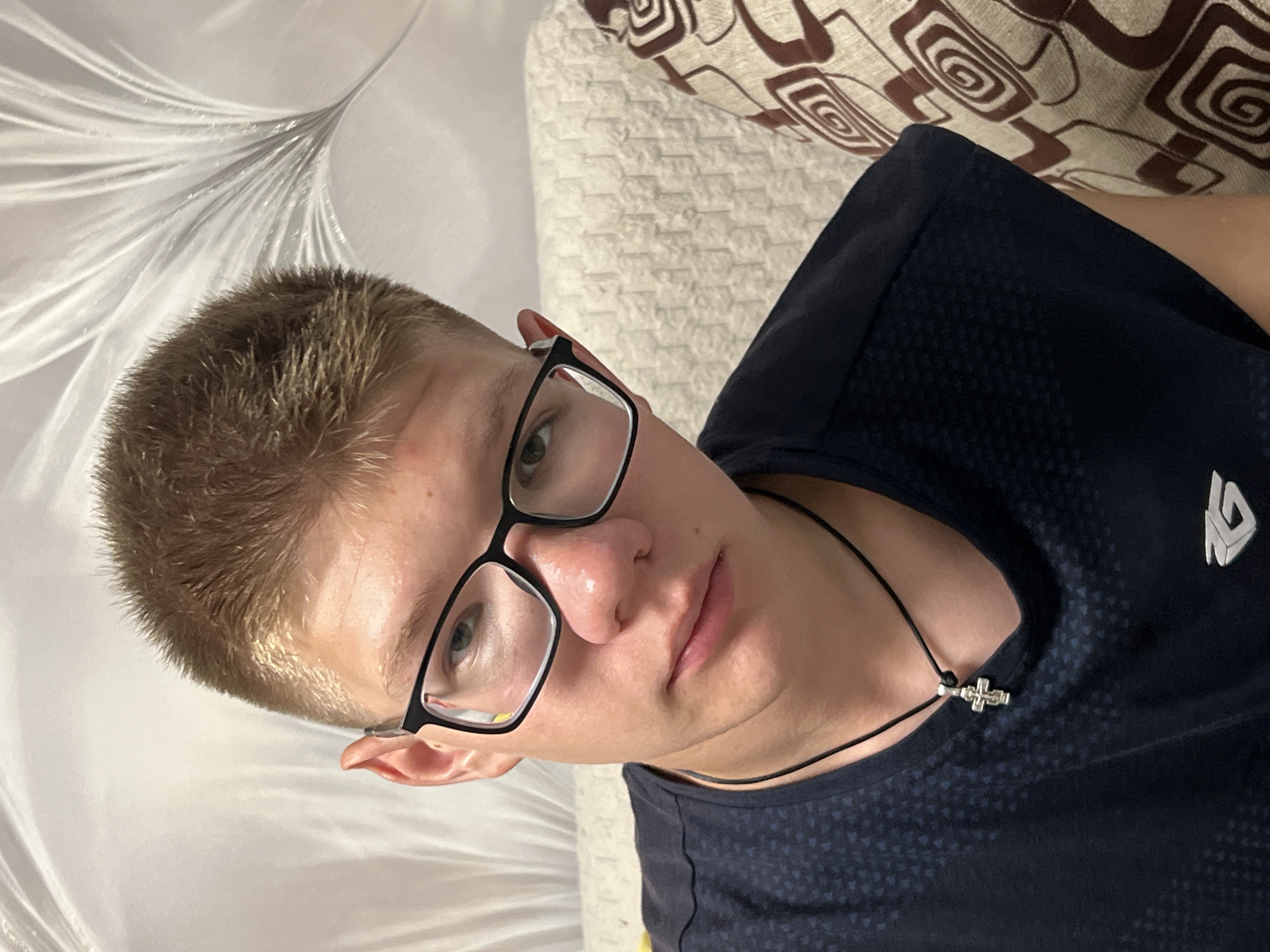 A young male with short, blonde hair and glasses is shown in a close-up portrait. He is wearing a dark blue t-shirt with a subtle pattern and a black cord necklace with a silver cross pendant. The background consists of a white curtain and a patterned sofa, suggesting an indoor residential setting. The image appears to be taken in Chaikovsky, Russia, based on the provided context.