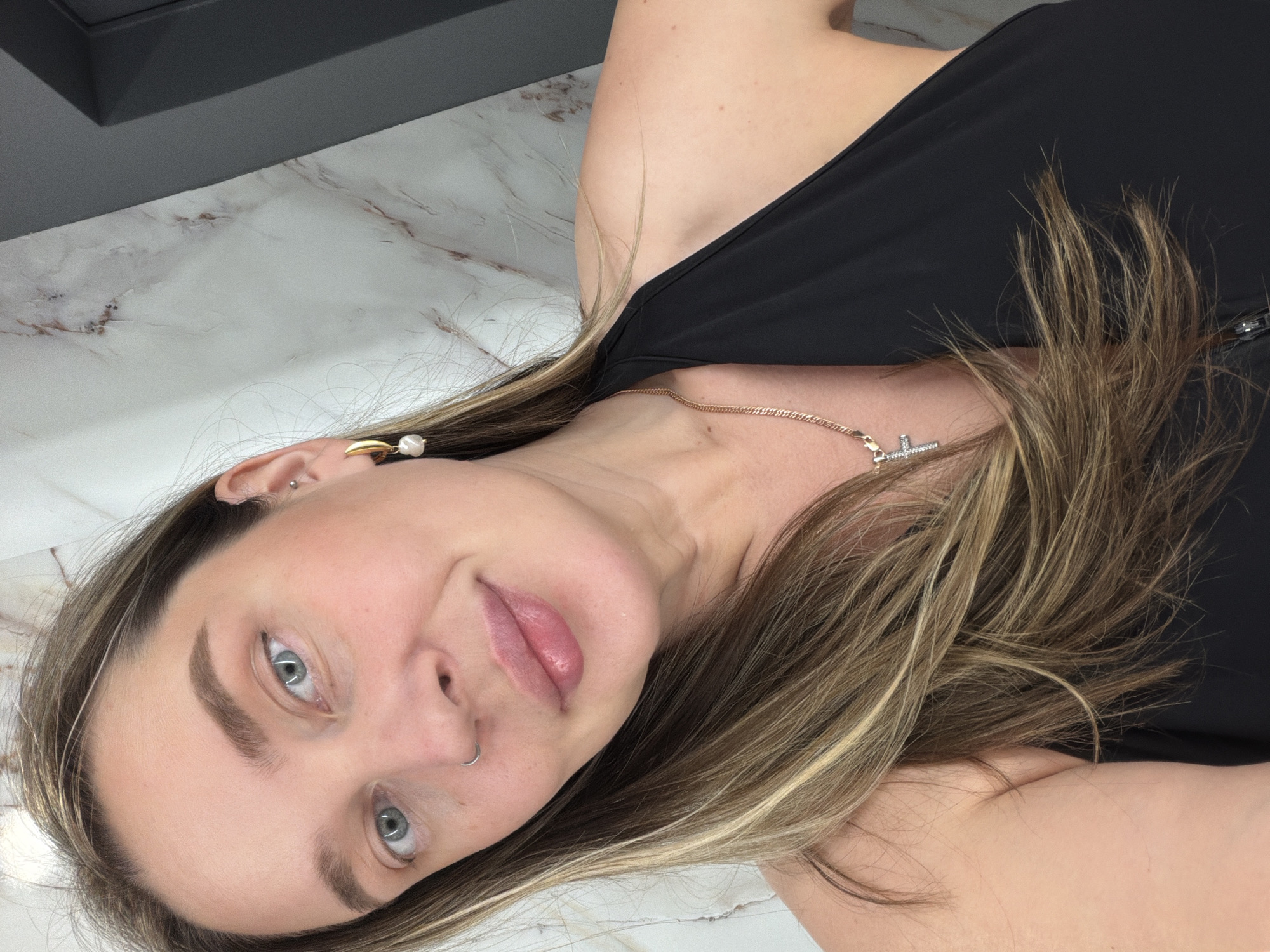 A selfie-style image depicts a woman with fair skin, light brown hair with blonde highlights, and blue eyes. She is wearing a black sleeveless top and a gold necklace with a small cross pendant. She has a piercing in her left nostril and two earrings in her left ear. The background consists of a white marble surface and a dark gray object in the upper left corner. The photo appears to be taken indoors. There is no evidence of Moscow, Russia.