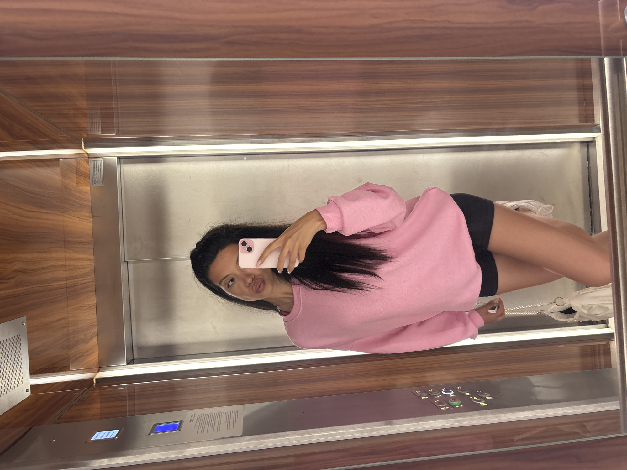 A young woman with long, dark hair, wearing a pink sweatshirt and black shorts, is taking a mirror selfie in an elevator. She is holding a pink phone in her right hand and has her lips pursed. A white purse with a pearl strap is hanging from her left arm. The elevator has wood-paneled walls and a stainless steel door. The background shows the interior of the elevator, including a control panel with buttons. The overall scene is a casual, everyday moment captured in what appears to be a modern building. The specific location in Sofia, Bulgaria, cannot be determined from the visual information provided.