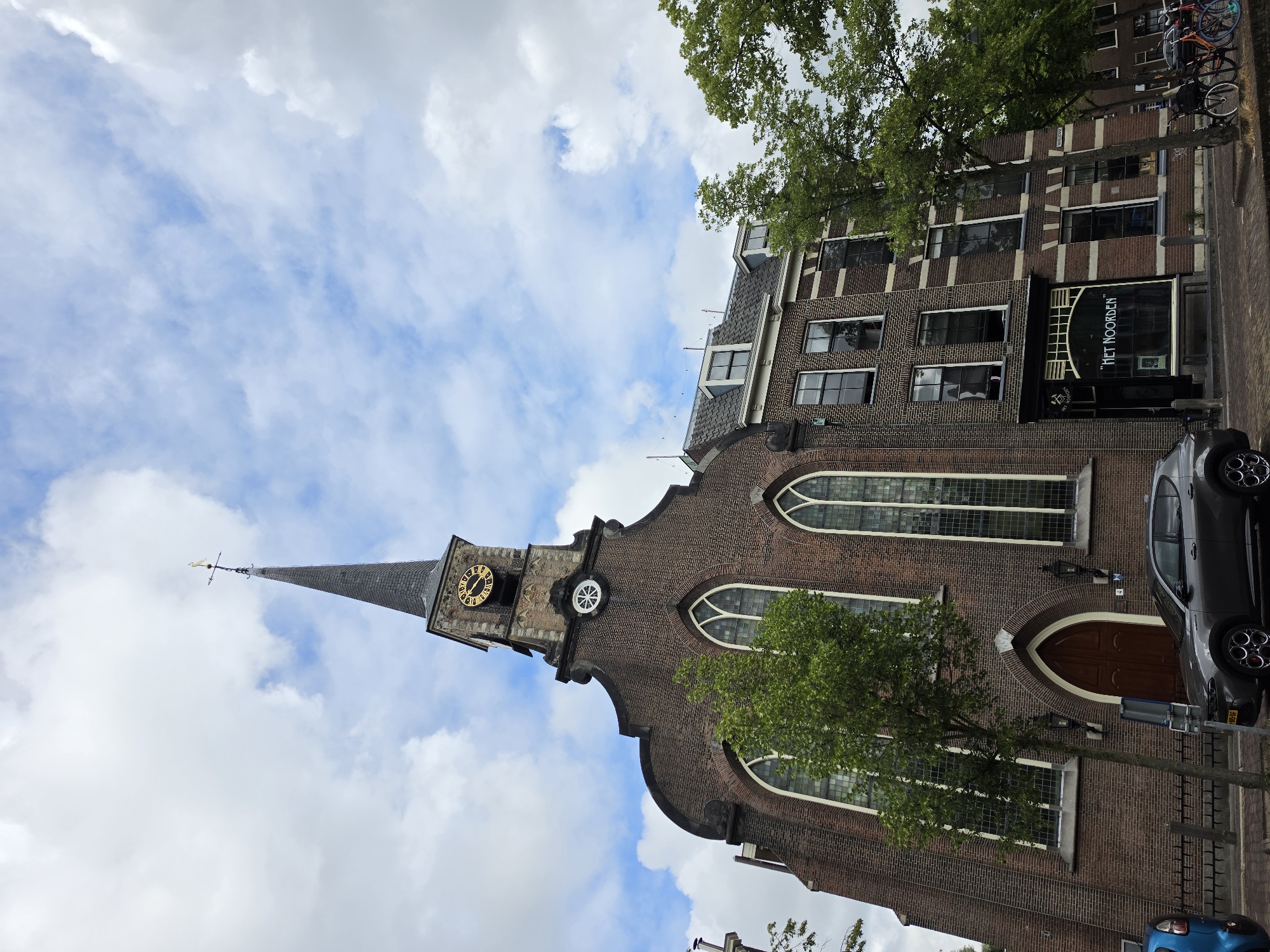 The image shows the exterior of a church and adjacent buildings in Delft, Netherlands. The church has a prominent spire with a clock and a weather vane. It is constructed of red brick and features arched windows. Next to the church, there is a multi-story building with a shopfront that has the text "HET NOORDEN" displayed. Trees are visible in front of both the church and the adjacent building. A dark grey car is parked on the street in front of the church, and several bicycles are parked near the adjacent building. The sky is partly cloudy.
