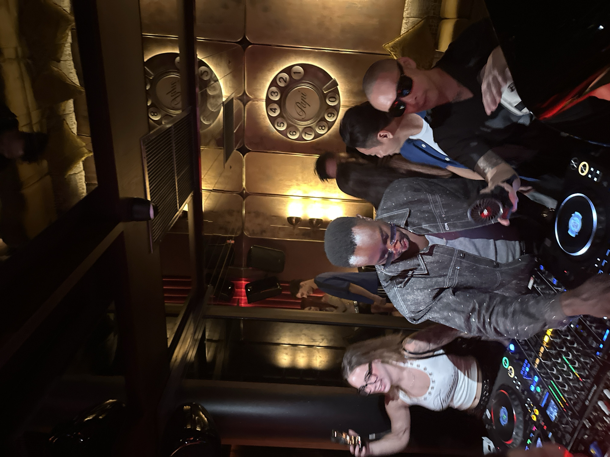 The image shows a DJ booth with two DJs in a dimly lit club. The woman in the foreground wears a white top and glasses, holding a smartphone. The man beside her wears a grey patterned jacket. Patrons surround the DJs and equipment. The walls are decorated with lighted rotary telephone style panels. This appears to be the location Papi's in Thủ Đức, Vietnam.