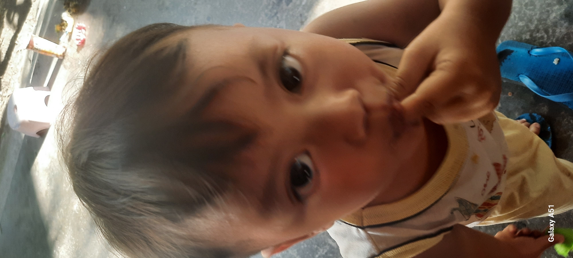The image shows a close-up, overhead view of a young child, possibly a toddler, wearing a light-colored, sleeveless shirt with a yellow trim and a printed design. The child is outdoors, standing on a concrete surface, and is in the process of putting a finger in their mouth. In the background, a light-colored stool and some scattered objects are visible on the ground. A blue flip-flop is also present near the child's feet. The scene is illuminated by sunlight, creating shadows on the ground. The image is tagged with "Galaxy A51," indicating it was taken with a Samsung Galaxy A51 smartphone. The location is identified as Ploso, Indonesia.