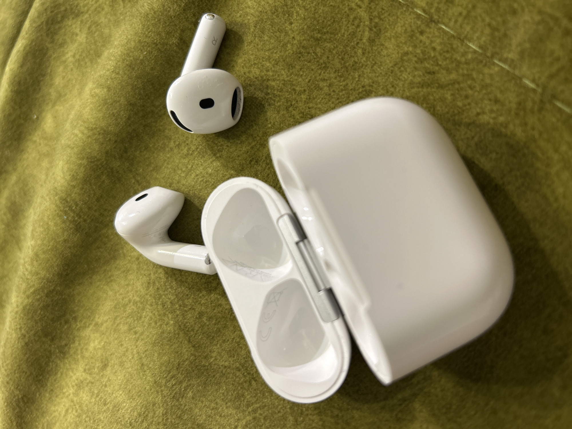 A white Apple AirPods Pro charging case is open on its side on a green textured surface. Inside the case, the white earbuds are not visible. To the left of the case, a single white earbud is partially visible, oriented with its stem pointing upwards and its speaker grill facing left. To the right of the case, another single white earbud is visible, lying on its side with the stem pointing towards the right. This earbud has a black circular speaker grill and the letter 'R' is visible on its stem. The green surface appears to be a soft fabric, possibly a blanket or a couch. The lighting suggests an indoor environment.