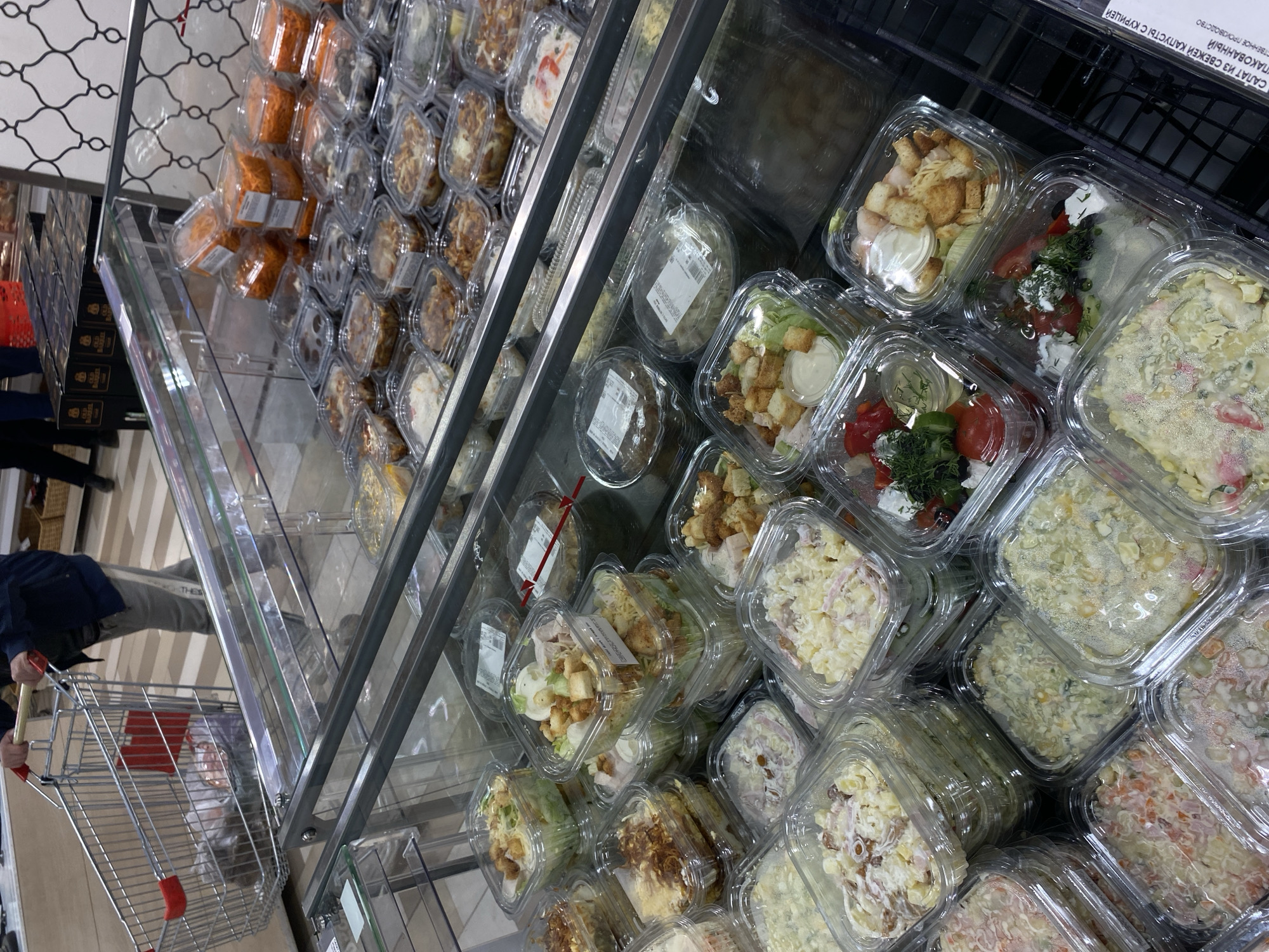 The image shows a refrigerated display case filled with various pre-packaged salads and other prepared food items. The items are arranged on multiple shelves within the case. The foreground includes a shopping cart. A person is visible pushing the cart, with the focus being on the display case's contents. The image appears to be taken inside a supermarket or grocery store. According to the text in the upper right corner, this is a supermarket in Kazan, Russia.