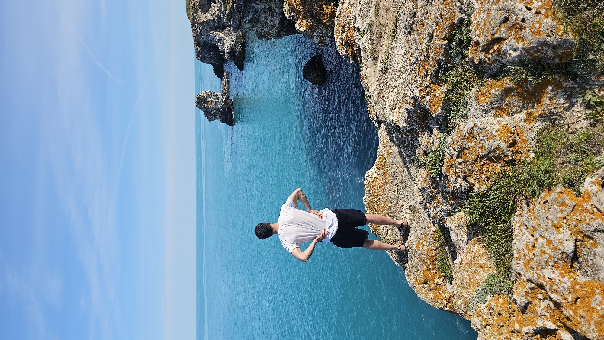 A man stands on a rocky cliff overlooking the sea in Tyulenovo, Bulgaria. He is facing the water, with his hands on his lower back. He is wearing a white t-shirt and dark shorts. The sea is turquoise blue, and the sky is light blue. There are rock formations in the sea near the cliff. The cliff is covered with orange moss and grass.