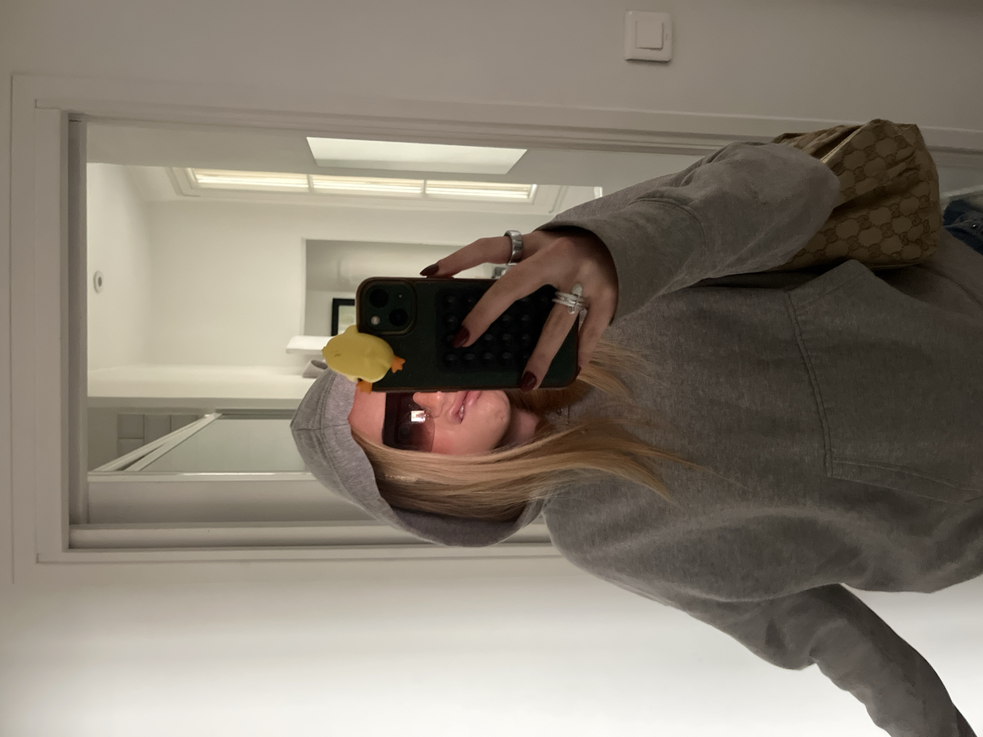 A woman is taking a selfie indoors, presumably in a mirror reflecting an apartment interior with white walls, a window with a skylight, and a hallway leading to other rooms. She is wearing a grey hoodie, sunglasses, and several rings on her fingers. She is holding a smartphone with a yellow duck accessory attached to it. A handbag is visible on her shoulder. The location cannot be determined to be Paris, France from this image.