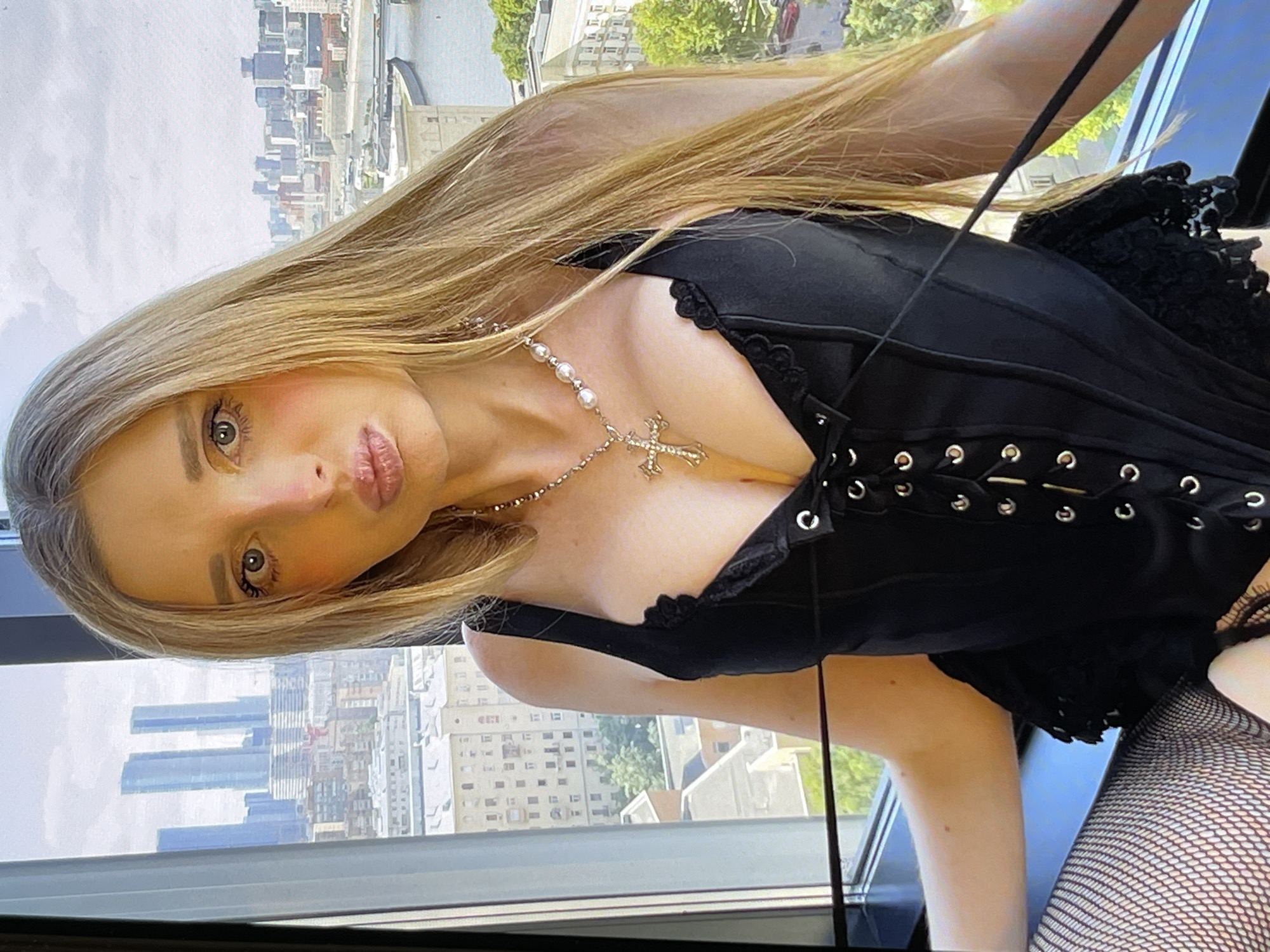 A medium shot shows a young woman with long blonde hair, wearing a black lace-up corset and fishnet stockings, posing in front of a window overlooking a city skyline. The woman has her lips pursed and is looking directly at the camera. Her eyes are wide and her makeup includes pink blush on her cheeks. She is wearing a silver necklace with a cross pendant and pearls. The background shows a cityscape with tall buildings and a body of water. The lighting is bright, suggesting it is daytime.