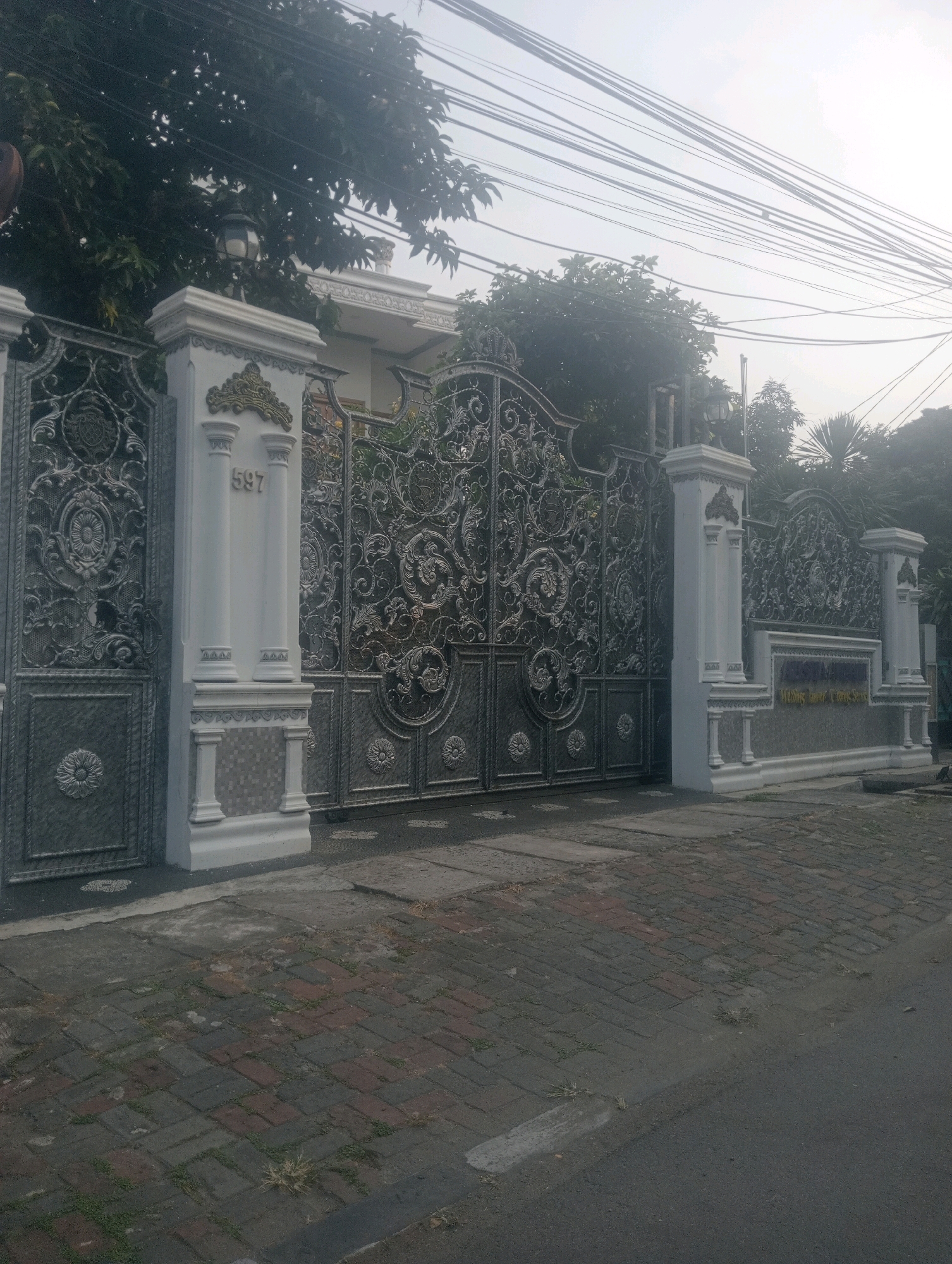 This is a medium-shot, eye-level, outdoor photograph of an ornate metal gate and its accompanying stone pillars, located on a street in East Jakarta, Indonesia. The gate is made of intricately designed metalwork featuring floral and scrolling patterns, and it appears to be a double gate. To the left of the gate, there is a white stone pillar with the number "597" embossed on it. To the right of the gate, there is a long, low stone wall with a sign that is partially obscured by glare but appears to have text on it. The street in front of the gate is paved with bricks, and to the right, there is asphalt. Trees with dense foliage are visible behind the gate, and power lines are strung across the sky. The overall lighting suggests it is either early morning or late afternoon, with a slightly hazy atmosphere. There are no people visible in the image. The scene depicts the entrance to a property, likely a residence or a business, characterized by its elaborate and well-maintained gate.