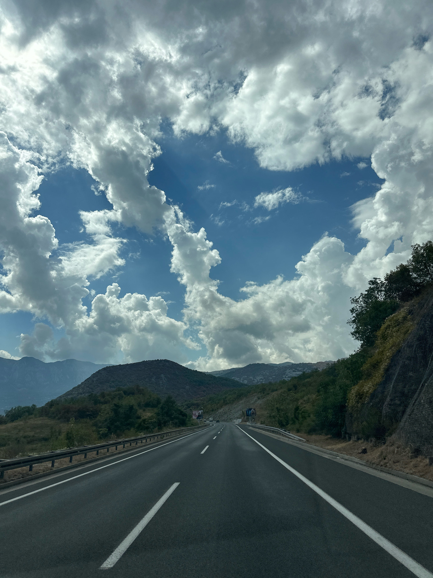 The image shows a road in Sotonići, Montenegro, with mountains and a cloudy sky in the background. The road is asphalt and has white lines marking the lanes. There are guardrails on the side of the road, and some vegetation and rocky slopes. The sky is filled with dramatic clouds, with patches of blue sky visible. The sun is shining through the clouds, creating rays of light. There are no people or vehicles visible in the image.