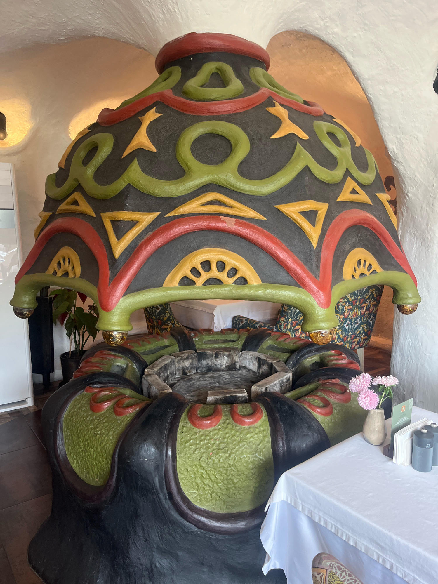 The image shows a large, ornate, sculptural fireplace that resembles a giant, colorful urn or bell. It is painted in dark gray, red, green, and yellow, with intricate swirling patterns and geometric shapes. The fireplace is centrally located in what appears to be a restaurant or indoor seating area. In the background, the walls have a rough, textured finish and are illuminated by warm lighting, creating a cozy atmosphere. To the left of the fireplace, a tall plant in a pot is visible, along with a glimpse of a white refrigerator. To the right, a white-covered table is set with a vase of pink flowers, napkins, and salt and pepper shakers, suggesting it is ready for diners. The overall impression is of a unique and artistic interior design. The specific location in Krasnaya Polyana, Russia, is not discernible from the visual information provided.