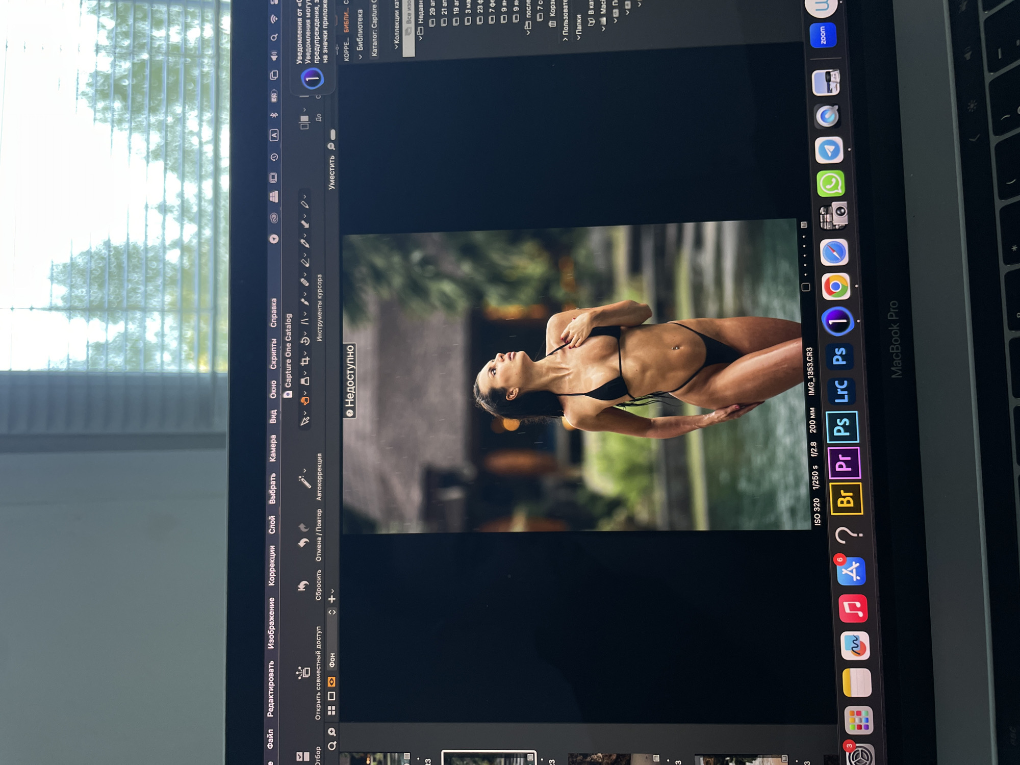 The image is a photograph of a laptop screen. The screen displays a photo editing software called Capture One. The photo being edited depicts a woman in a black bikini standing in an outdoor setting with lush green foliage, appearing to be in a tropical or subtropical location. The software interface shows various editing tools and parameters, including ISO 320, 1/250s, f/2.8, and 200mm. The laptop is a MacBook Pro, evidenced by the branding on the keyboard deck. The operating system's dock is visible on the right side, displaying various icons like Zoom, WhatsApp, and other Apple applications. A window blind is partially visible in the background behind the laptop. There is no indication the location is in Kazan, Russia.