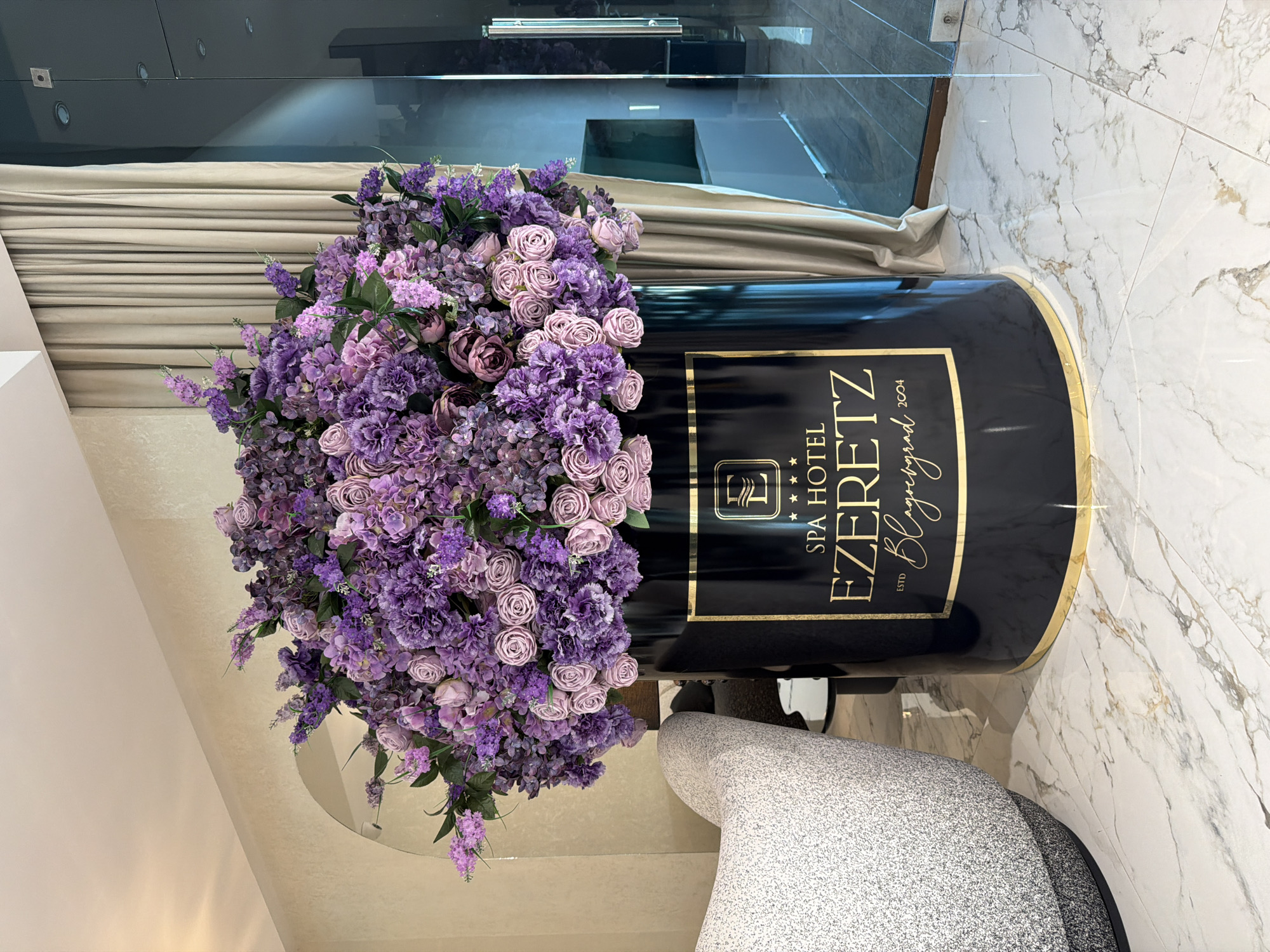 The image captures a floral arrangement inside the Spa Hotel Ezeretz in Blagoevgrad, Bulgaria. The arrangement consists of purple and pink flowers overflowing from a black container decorated with gold trim. The container prominently displays the hotel's name, "SPA HOTEL EZERETZ," along with "the Blagoevgrad" and "ESTD 2004." The arrangement stands on a light-colored floor with a marble pattern. Part of a gray chair is visible near the flower arrangement. The background includes beige curtains, and a glass wall or window. The orientation of the image is vertical.