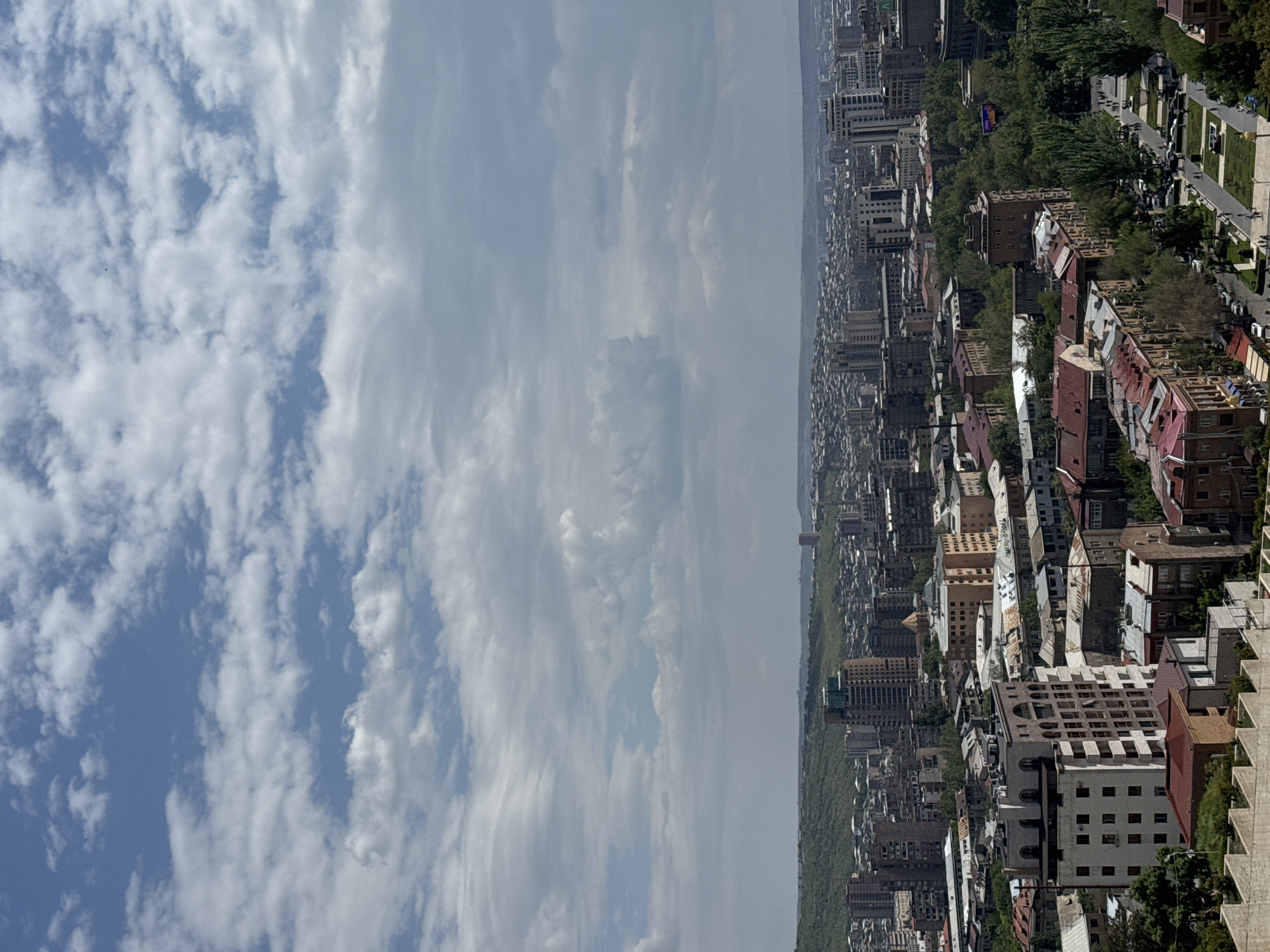 The image shows an elevated view of Yerevan, Armenia, on a day with a partly cloudy sky. The city stretches into the distance. Buildings of various heights and architectural styles are densely packed, interspersed with greenery. A prominent avenue lined with trees and pedestrian walkways is visible. The photograph is oriented vertically, with the sky occupying a large portion of the left side.