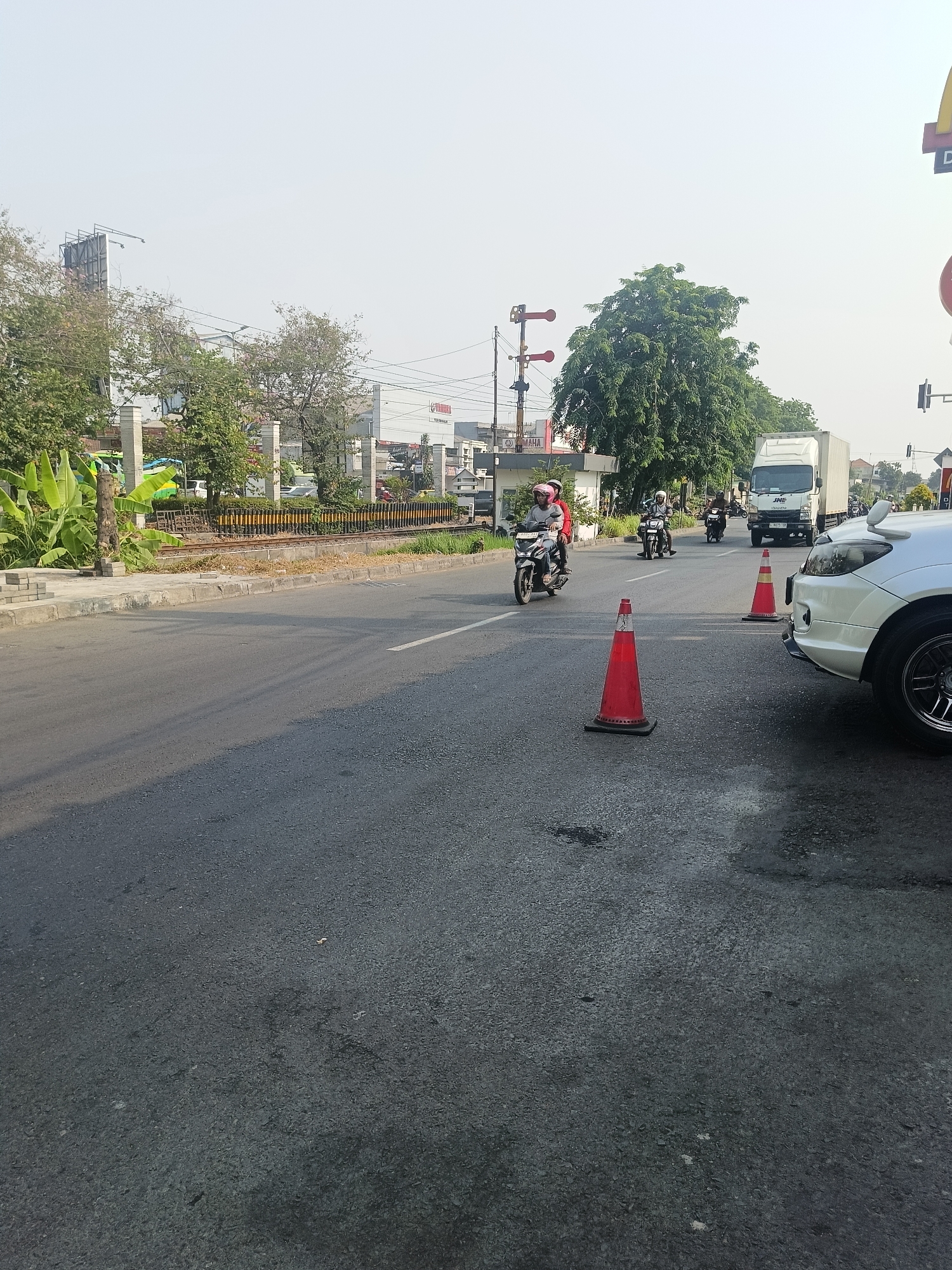 The image captures a daytime street scene in Calukan RT 1 RW 5 Keboansikep, Indonesia. A light-colored sedan is parked on the right side of the asphalt road. Two orange traffic cones are visible on the road, one in the foreground to the left of the sedan and another further back. Several motorcycles are on the road. One motorcycle with two riders is in the left lane, moving towards the viewer. Another motorcycle with a single rider is in the right lane, moving away from the viewer, followed by another motorcycle with a single rider. A white truck with the "JNE" logo and "ISUZU" visible on its front is in the right lane, also moving away from the viewer. Beyond the road, there is a railway track with yellow and black barriers. Trees, buildings, and utility poles are present in the background under a hazy sky.