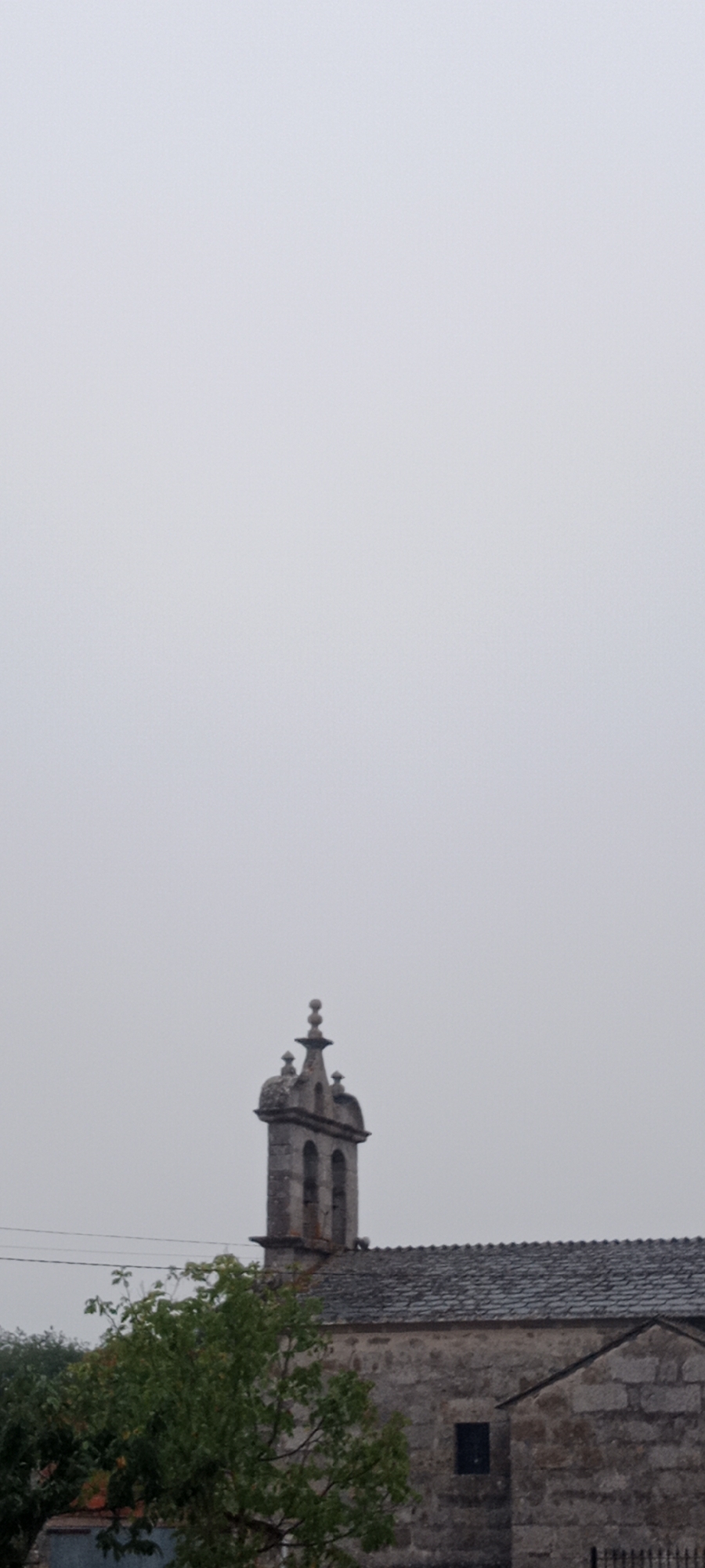The image shows a stone bell tower of a church against a light gray, overcast sky. The bell tower has a central arched opening and is topped with a decorative finial. To the right of the bell tower is the roof of the church, made of dark gray stone tiles. The church walls are also constructed of stone. In the foreground to the left, a lush green tree with dark branches partially obscures the view of the church. The overall scene suggests a potentially damp or misty day. This appears to be a church located in Adai, Spain.