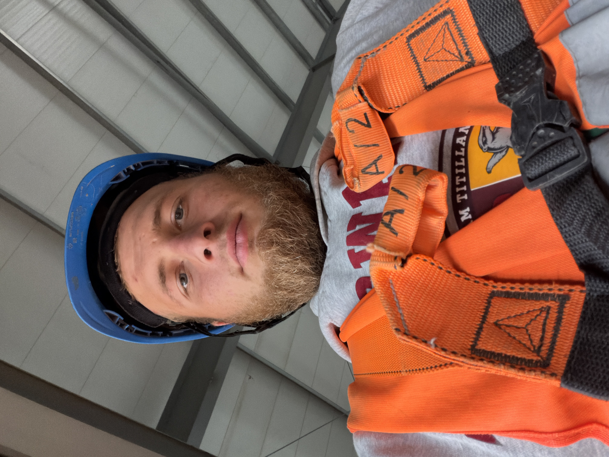 A man wearing a blue hard hat and an orange safety harness is taking a selfie in an industrial setting. The man has light skin, blue eyes, and a beard. He is wearing a grey hooded sweatshirt with a logo that appears to be a badger. The background consists of a corrugated metal ceiling and metal beams. This image was taken in Stróża, Poland.