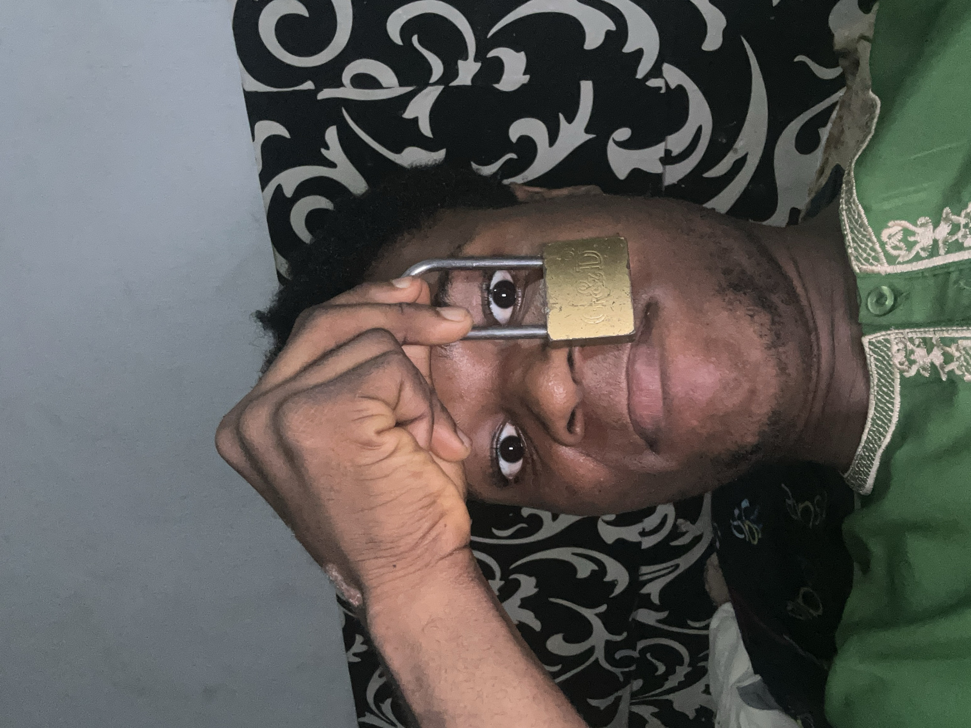 The image is a close-up selfie of a person holding a padlock over their right eye. The person has dark skin and is looking directly at the camera with their left eye open and wide. The padlock is gold-colored and appears to be slightly worn. The background is a patterned fabric, predominantly black and white, with a swirling design. The person is wearing a green garment with white embroidery. The lighting suggests an indoor environment, possibly at night. The image does not provide any specific information about the location in the city of Ihiagwa, Nigeria.